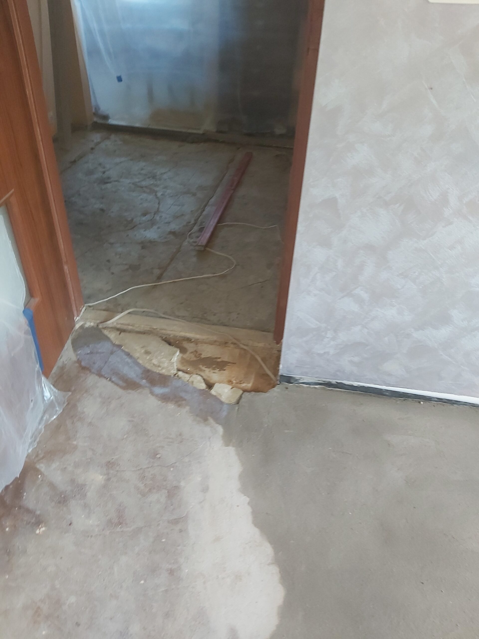 Widok progu drzwiowego z odsłoniętym betonowym podłożem i fragmentami starej warstwy, przygotowanymi pod wyrównanie posadzki.