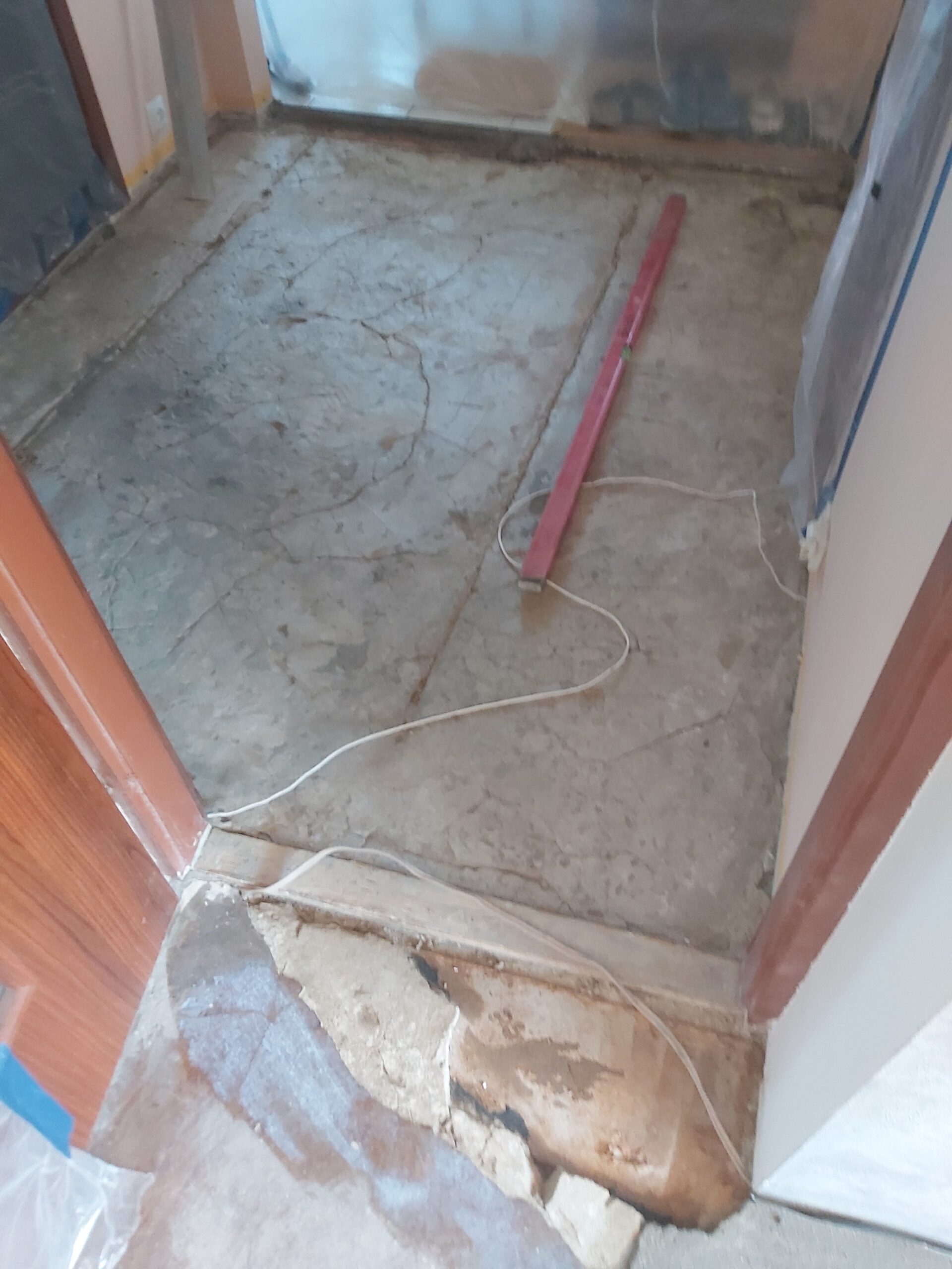 Odsłonięta betonowa podłoga w przejściu drzwiowym, z widocznymi pęknięciami oraz poziomicą i przewodem leżącymi na powierzchni.