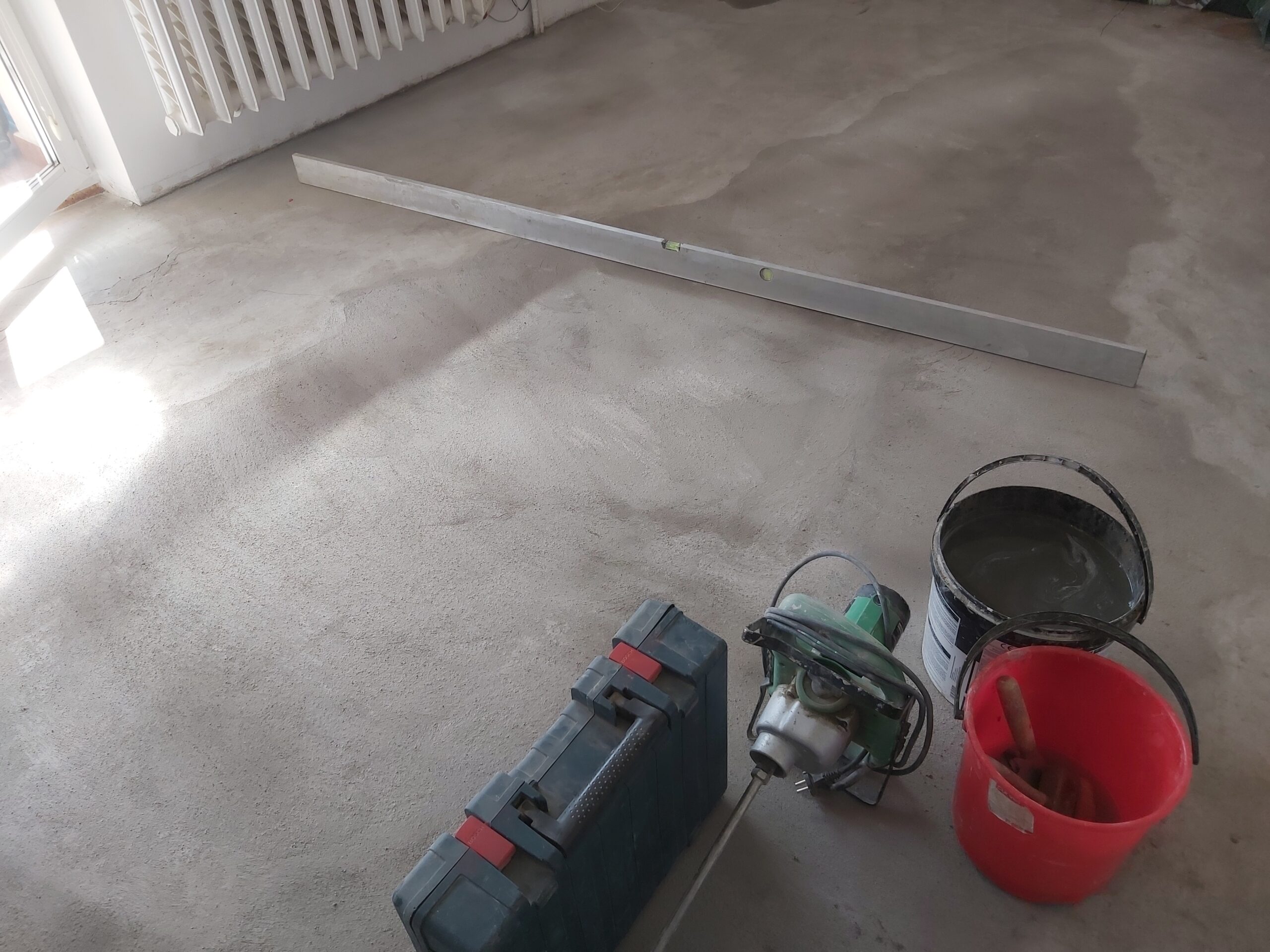 Posadzka cementowa z długą poziomicą oraz narzędziami budowlanymi przygotowanymi do dalszych prac wykończeniowych.