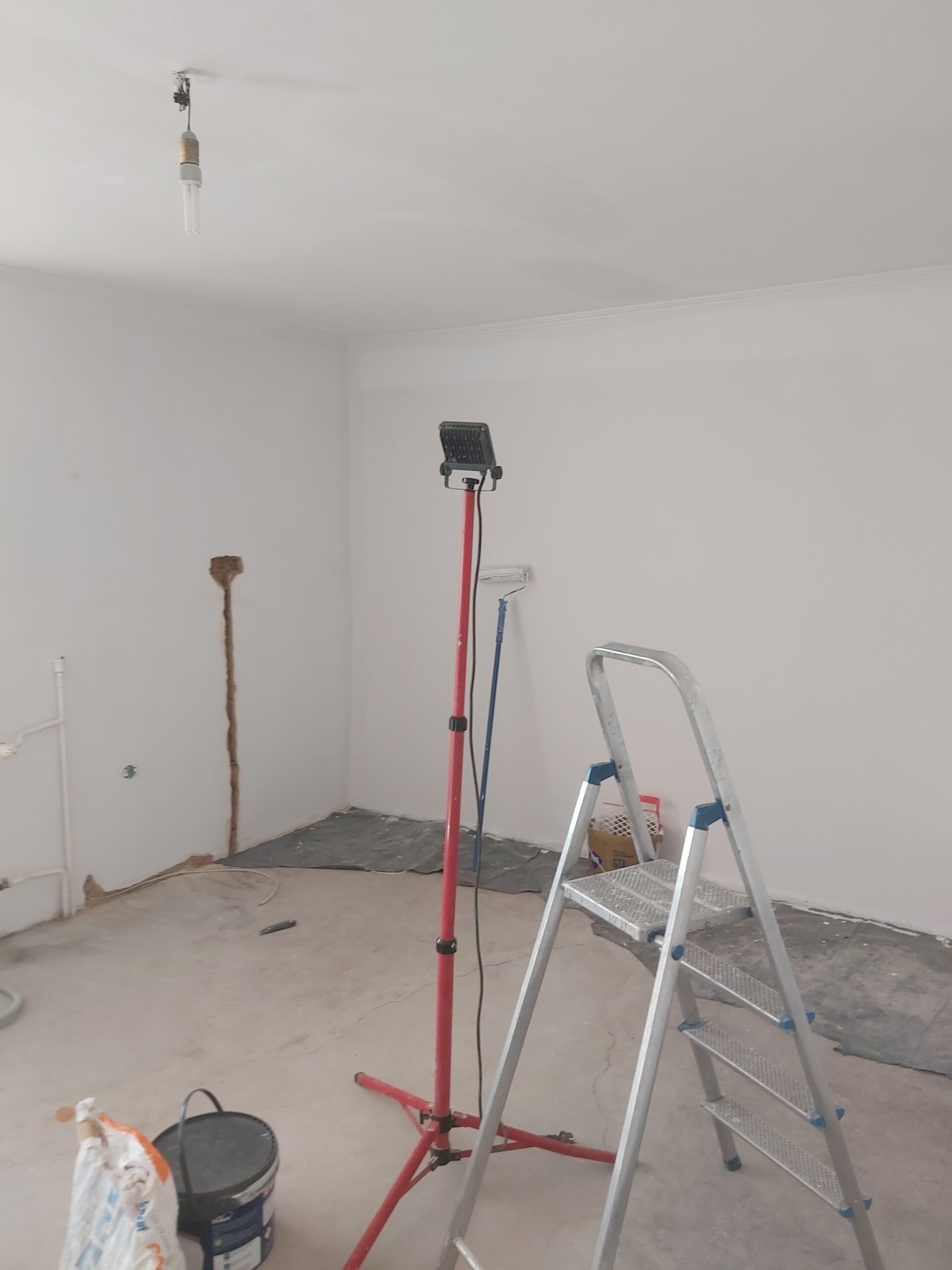 Wnętrze pomieszczenia w trakcie remontu z rozstawioną drabiną i lampą roboczą.