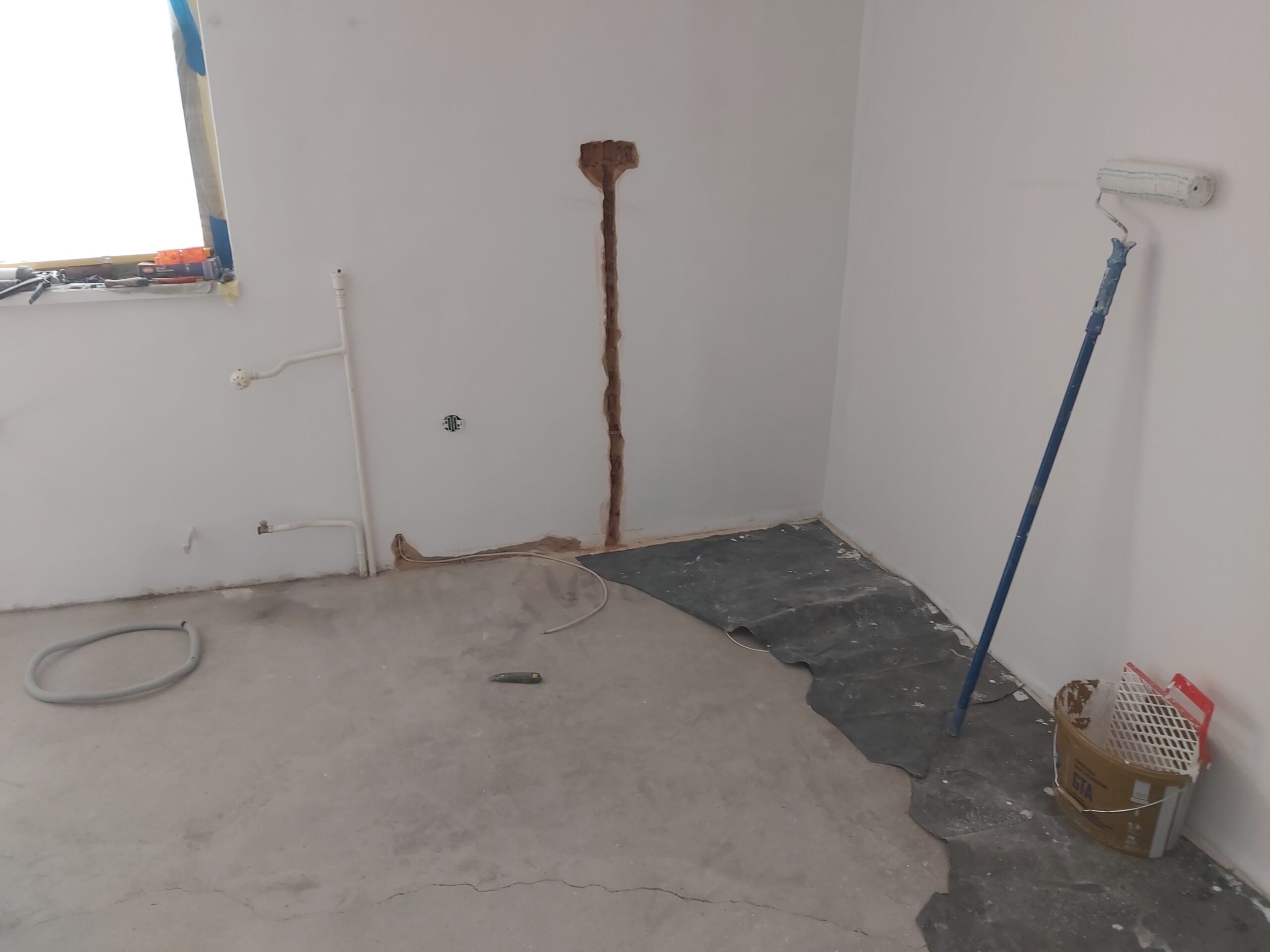 Pomieszczenie w trakcie remontu z odsłoniętą bruzdą instalacyjną w ścianie, fragmentem zabezpieczonej podłogi folią oraz wałkiem malarskim opartym o ścianę.