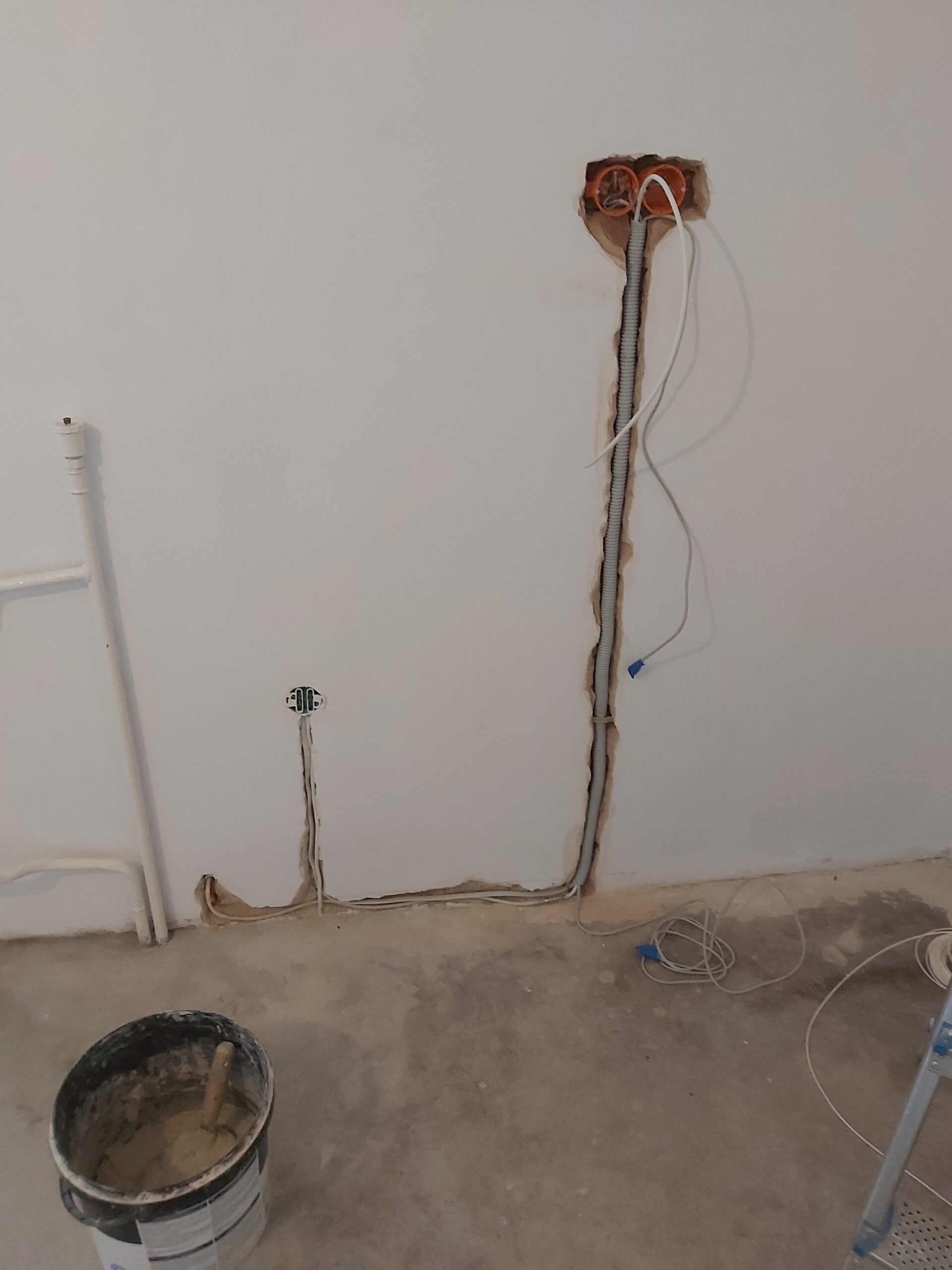Odsłonięta instalacja elektryczna w ścianie podczas remontu pomieszczenia, widoczne przewody, puszka elektryczna oraz wiadro z zaprawą na betonowej podłodze.
