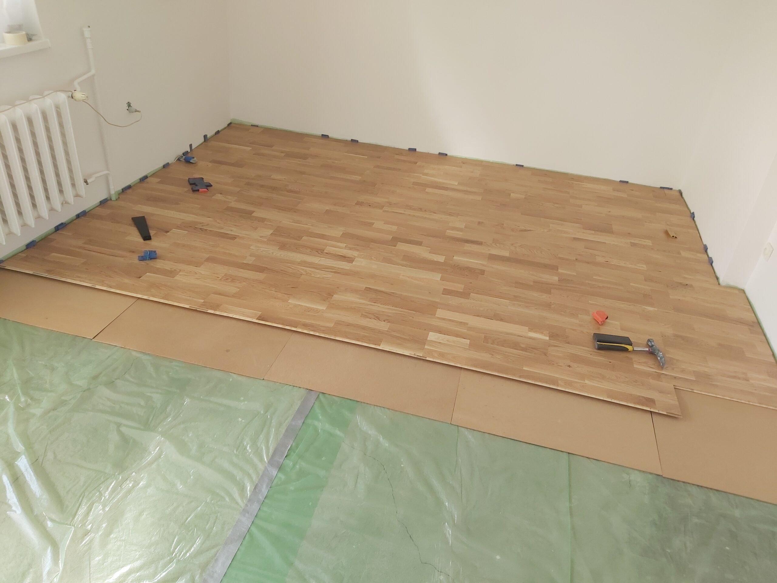 Pomieszczenie w trakcie montażu podłogi drewnianej z ułożonymi panelami, widocznymi narzędziami montażowymi oraz zabezpieczonym podłożem przed zabrudzeniem.