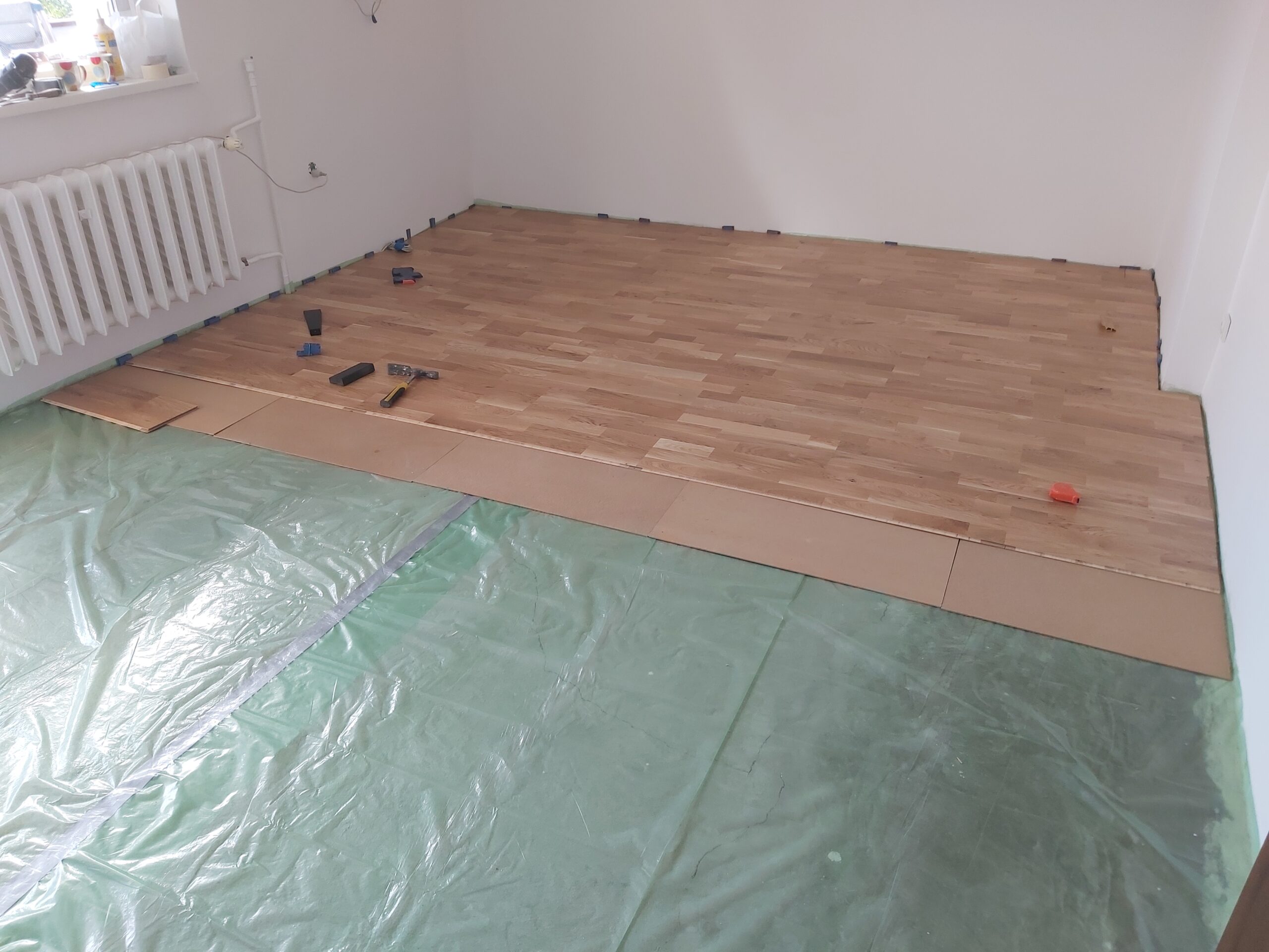 Drewniana podłoga w trakcie montażu, na podłożu leżą narzędzia, a część podłogi zabezpieczona jest folią ochronną.