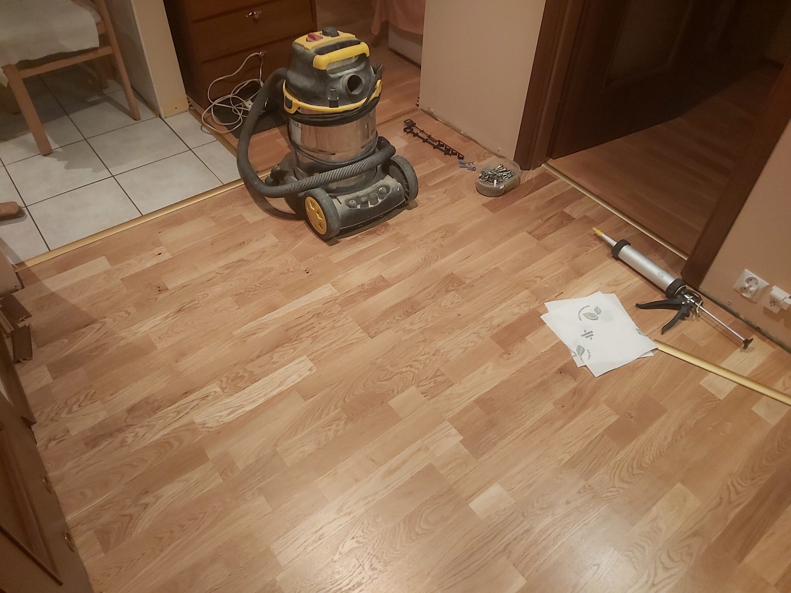 Drewniana podłoga w trakcie wykańczania, na podłodze stoi odkurzacz przemysłowy, leżą narzędzia montażowe i kartka z instrukcją.