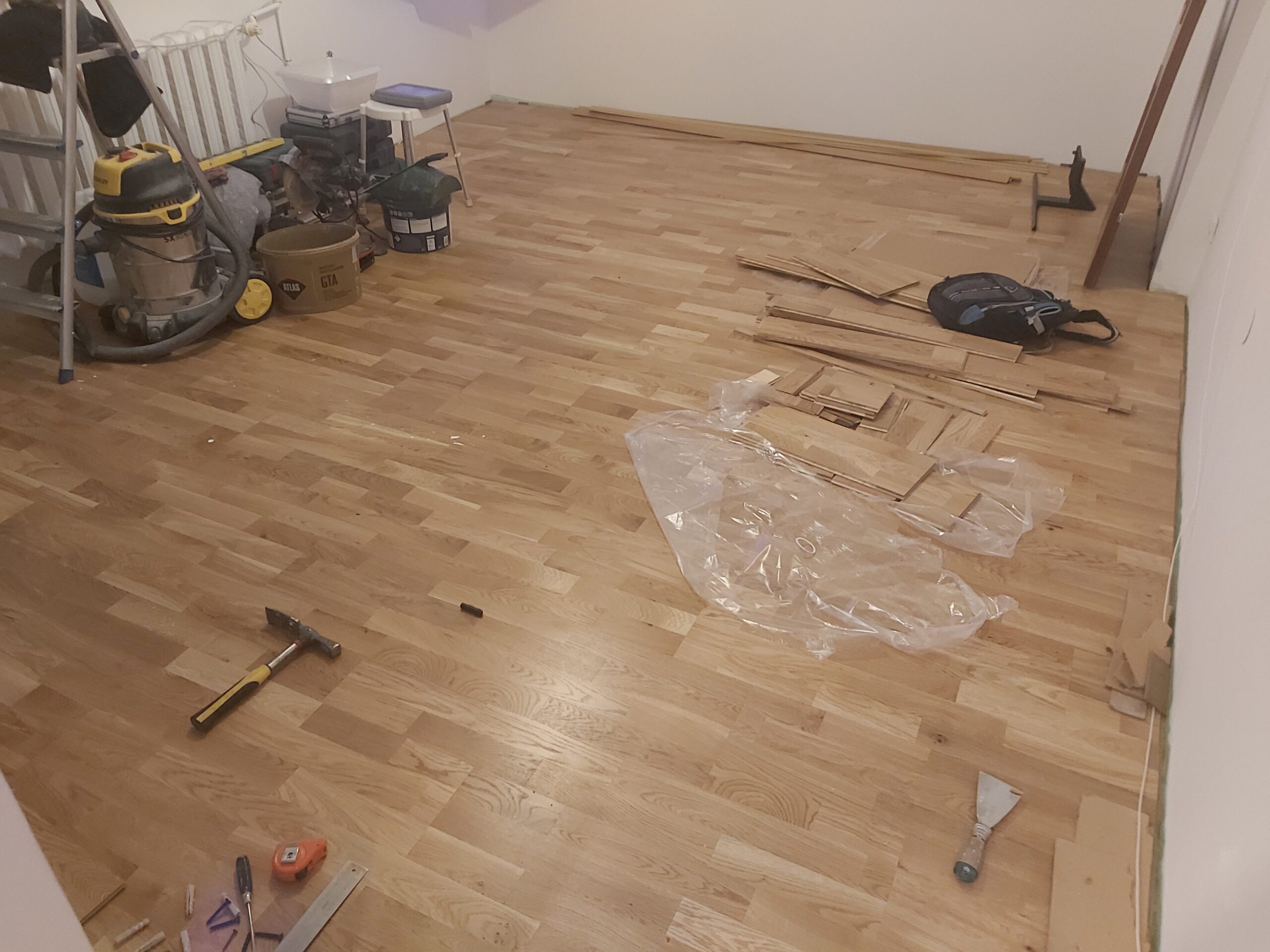 Drewniana podłoga podczas prac wykończeniowych, na podłodze leżą narzędzia, folie ochronne oraz pozostałe elementy montażowe.