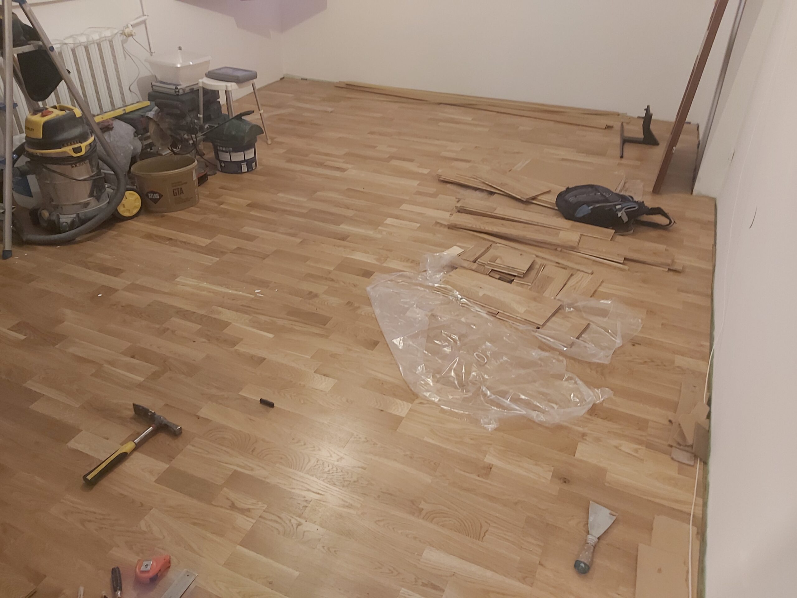 Drewniana podłoga w trakcie prac wykończeniowych, na powierzchni widoczne narzędzia, folie ochronne oraz elementy wykończeniowe przy ścianach.