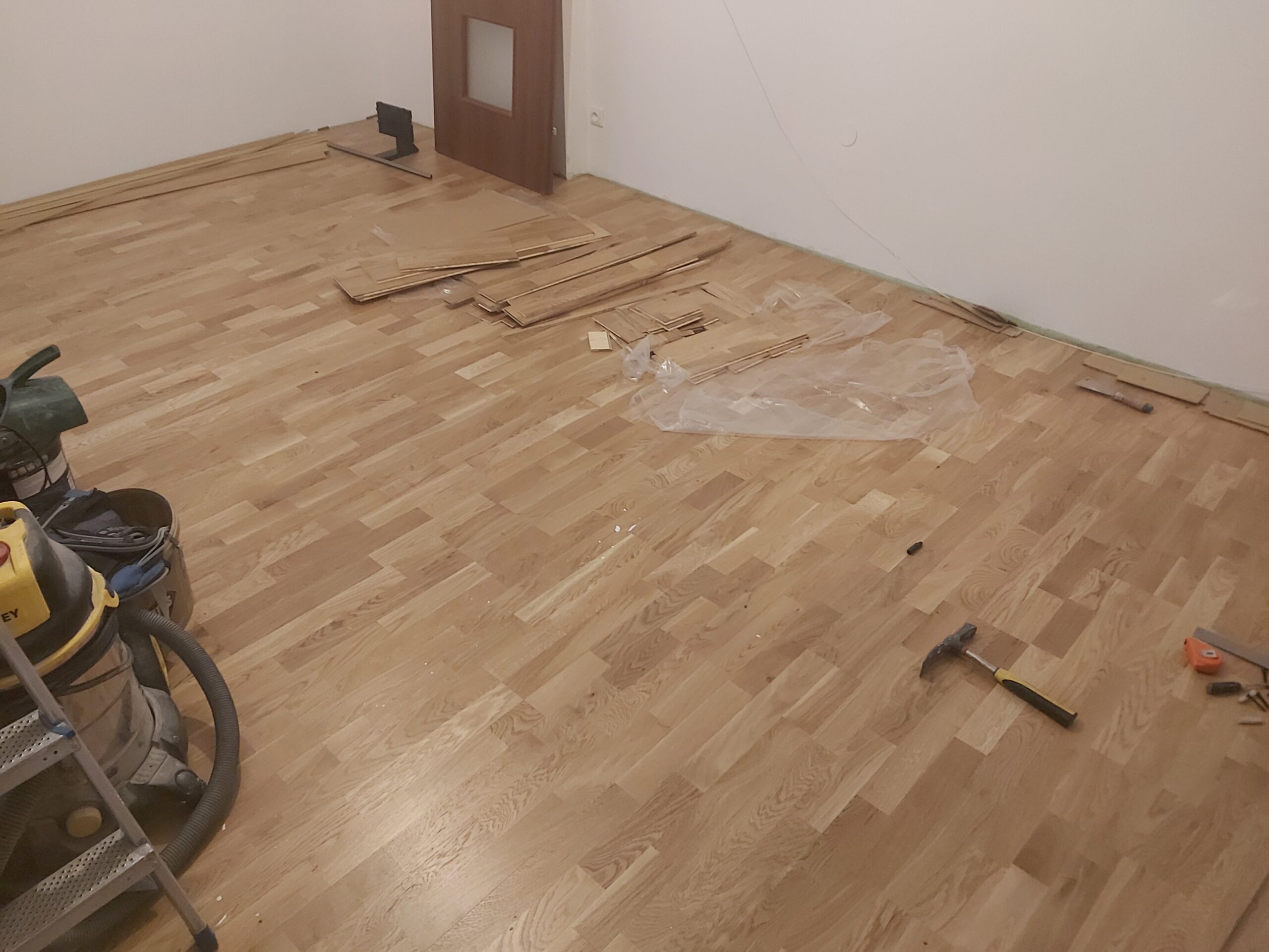 Drewniana podłoga w trakcie wykańczania, na powierzchni widoczne narzędzia, odkurzacz budowlany oraz pozostałości po montażu desek podłogowych.