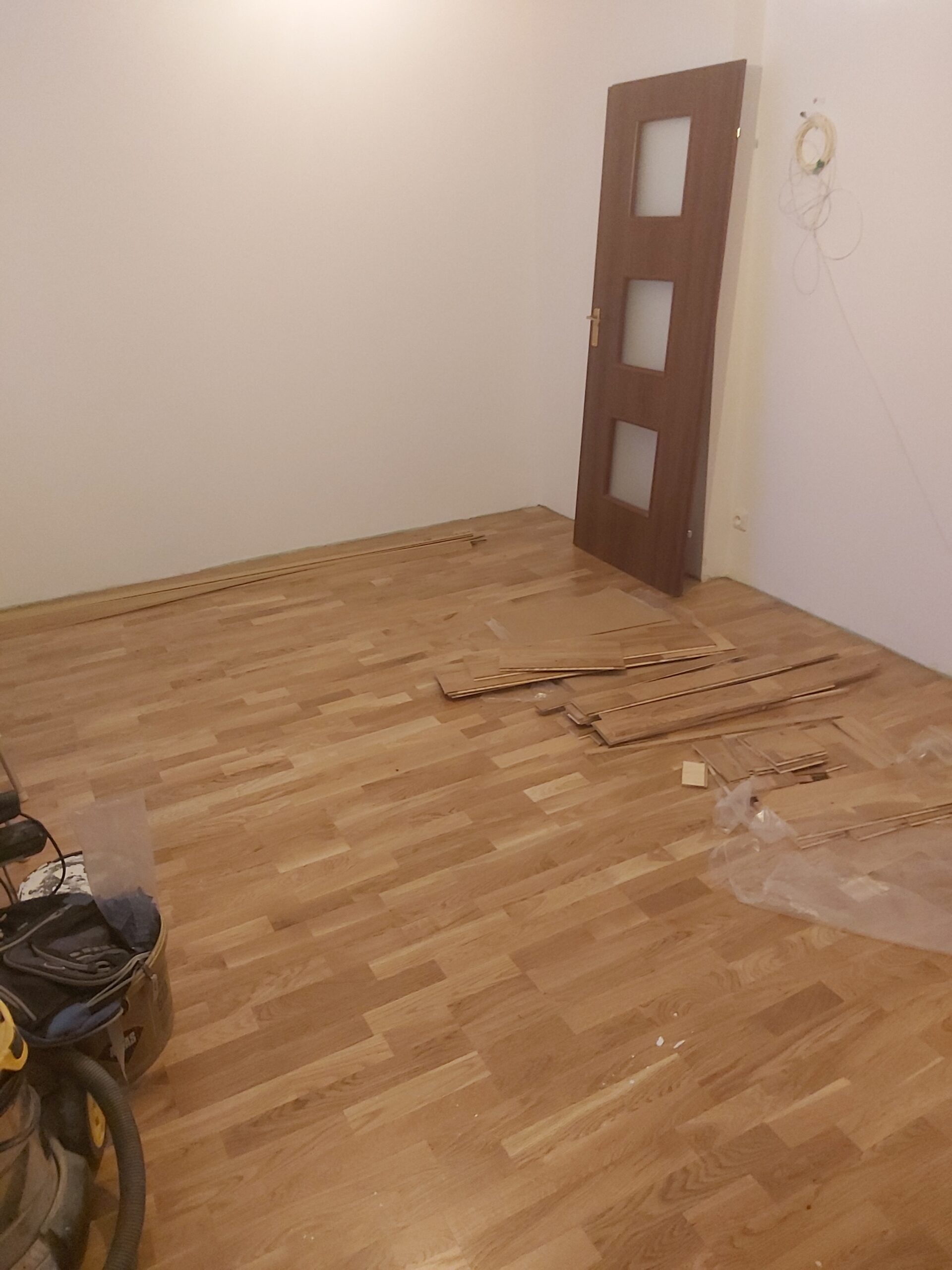 Pomieszczenie z ułożoną drewnianą podłogą, widoczne listwy i deski podłogowe przygotowane do montażu oraz drzwi oparte o ścianę.