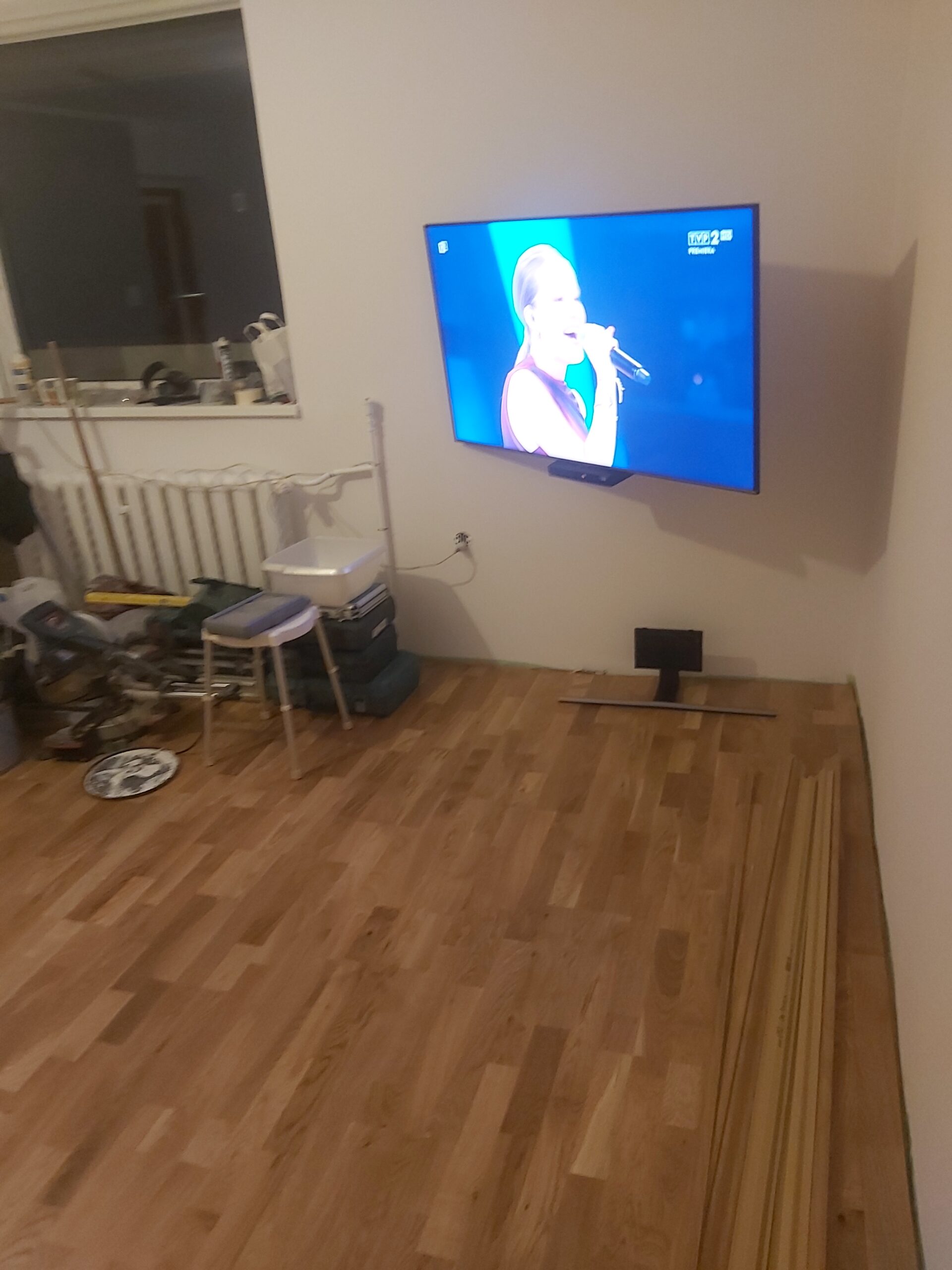 Telewizor zamocowany na ścianie w salonie podczas prac remontowych, wokół widoczne narzędzia, materiały budowlane oraz drewniana podłoga.