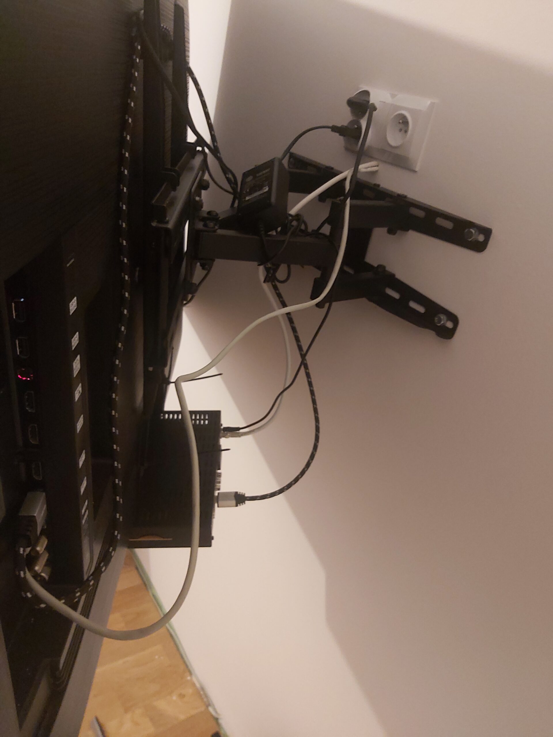 Tył telewizora zamontowanego na regulowanym uchwycie ściennym, widoczne przewody zasilające i sygnałowe podłączone do gniazdka oraz urządzeń multimedialnych.