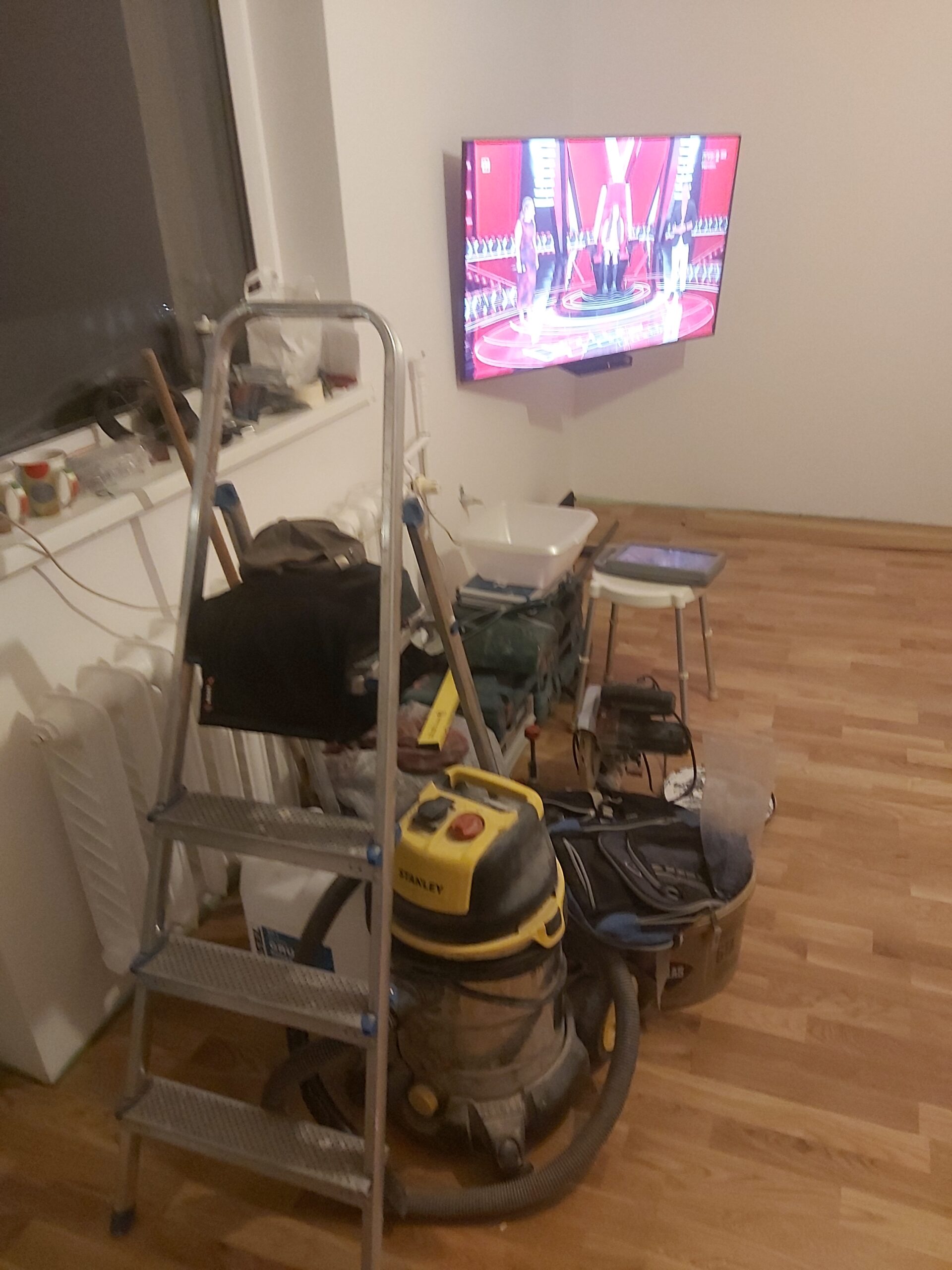 Wnętrze pokoju podczas prac remontowych – drabina, odkurzacz budowlany i narzędzia stoją przy ścianie, na której zamontowany jest włączony telewizor. Na podłodze panele, widoczne akcesoria i elementy wyposażenia w trakcie porządkowania.
