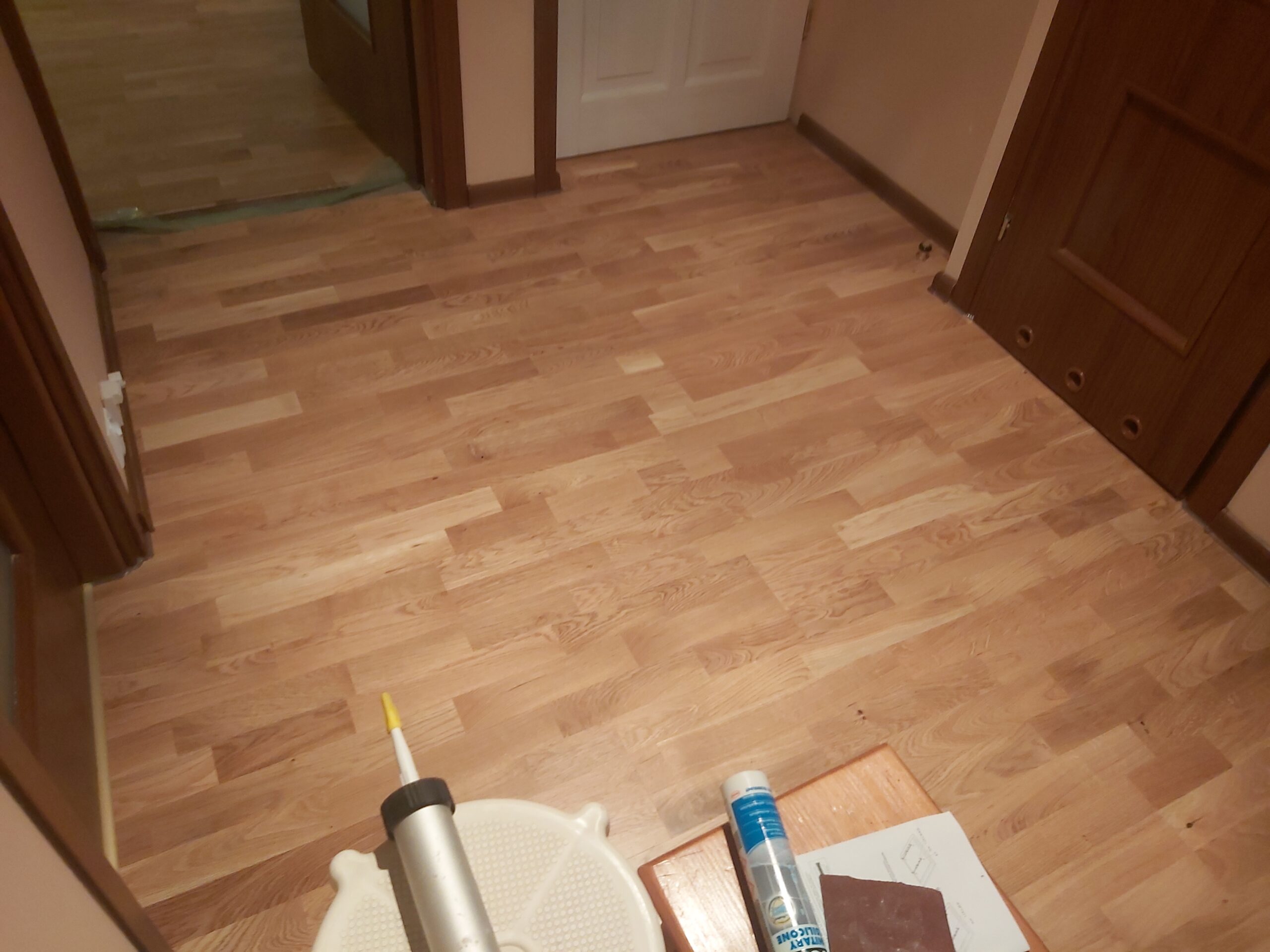 Widok korytarza z ułożoną drewnianą podłogą, widoczne drzwi do pomieszczeń i materiały montażowe na pierwszym planie.