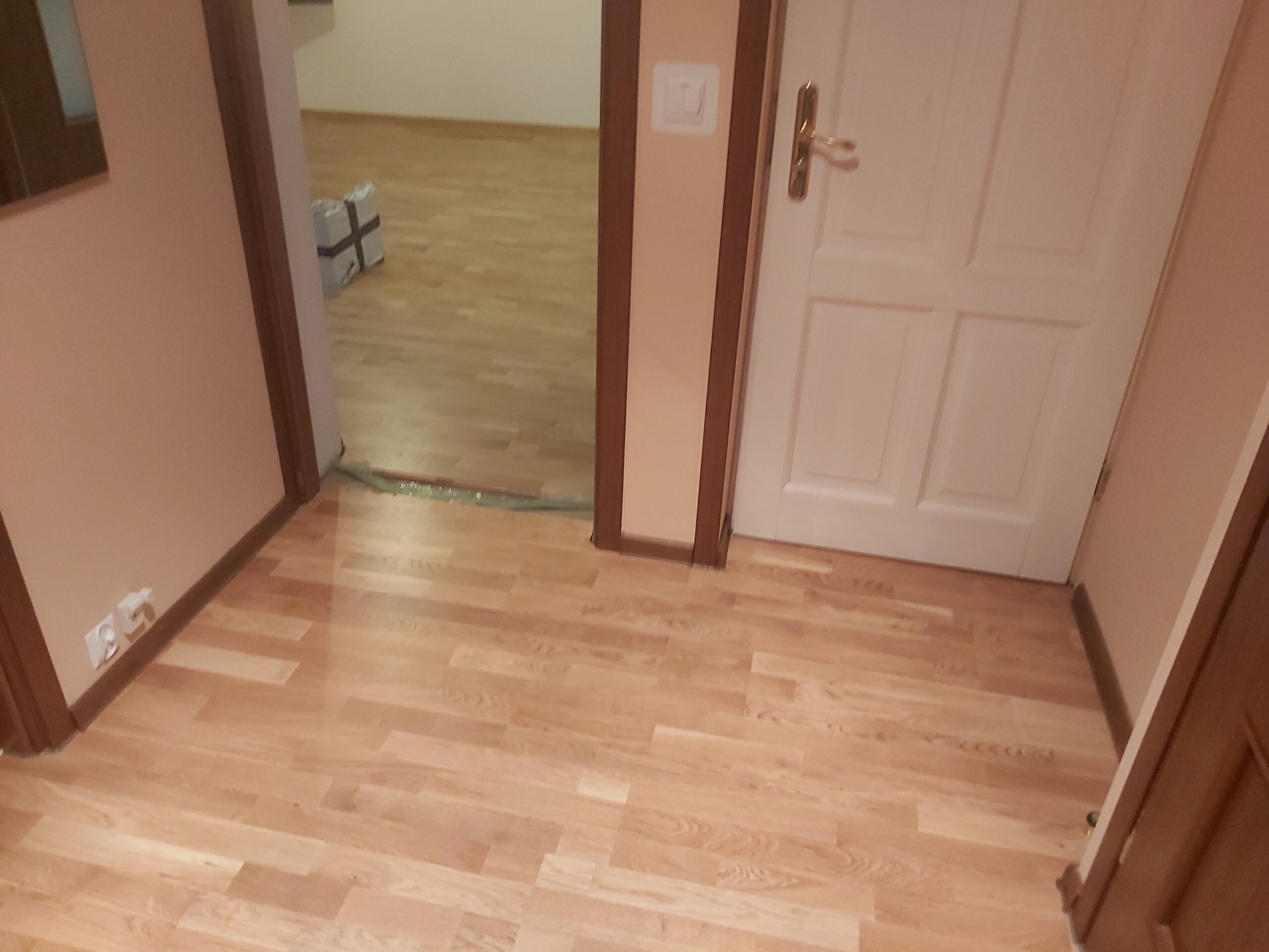 Widok korytarza z drewnianą podłogą i przejściem do sąsiedniego pokoju z taką samą podłogą.