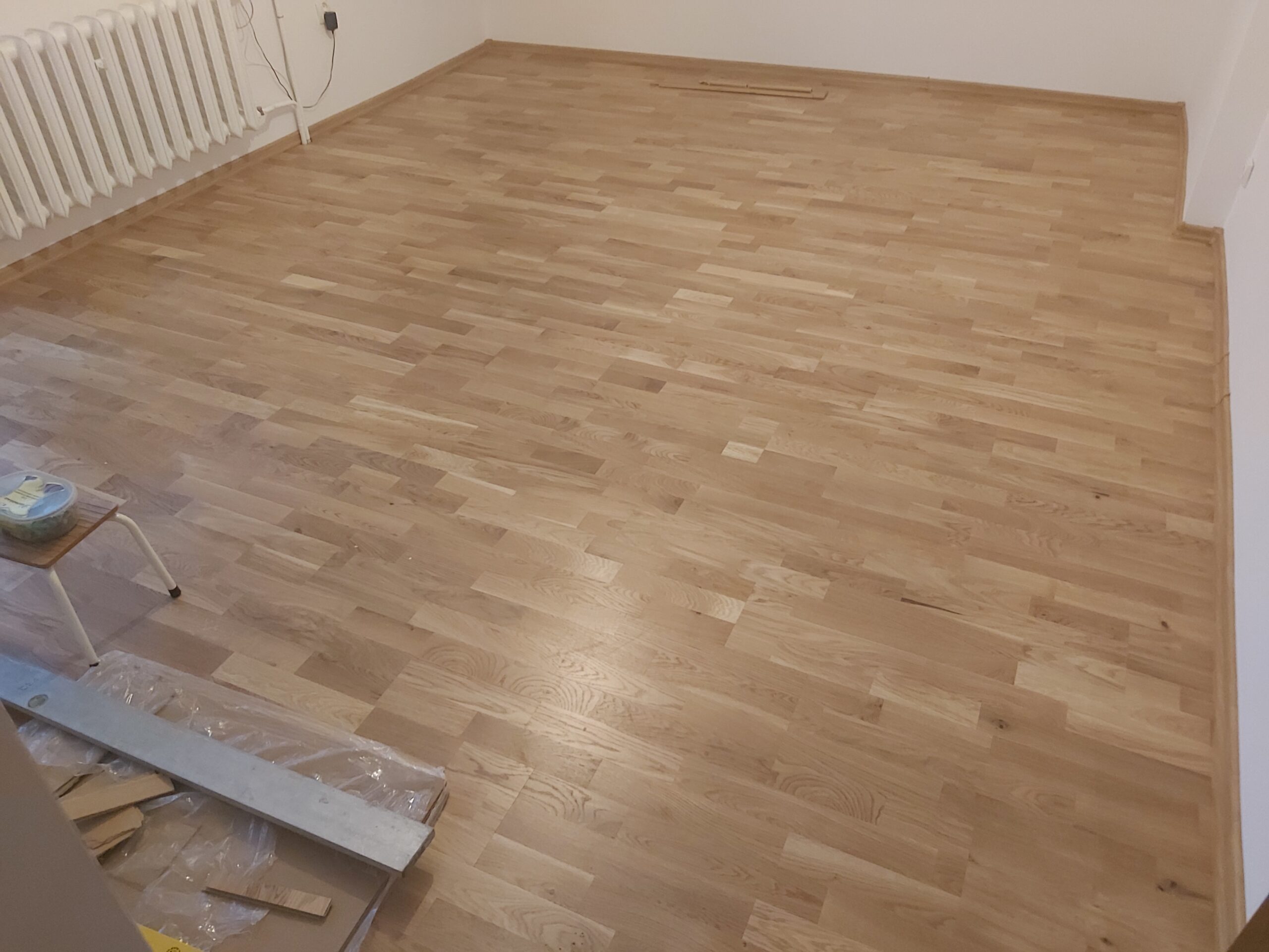 Widok na świeżo ułożoną drewnianą podłogę w pustym pomieszczeniu, z grzejnikiem przy ścianie.