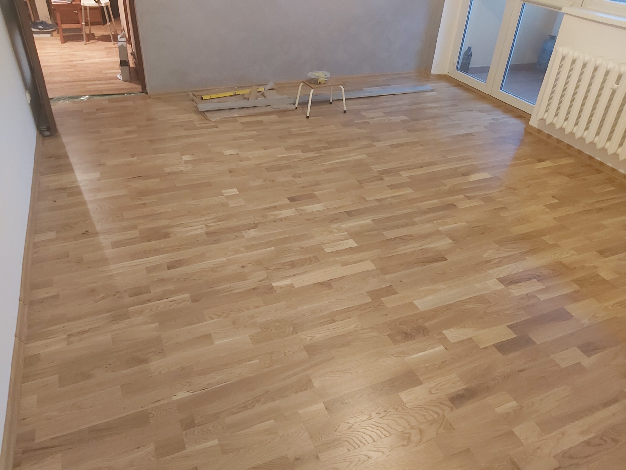 Wykończone pomieszczenie z ułożoną drewnianą podłogą, widok na grzejnik i drzwi balkonowe.