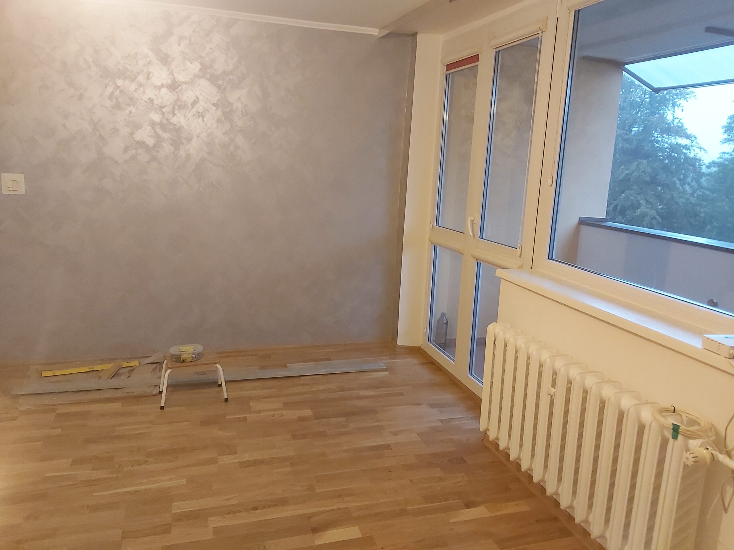 Pomieszczenie po remoncie z ułożoną podłogą, wykończonymi ścianami oraz widokiem na okna i grzejnik.