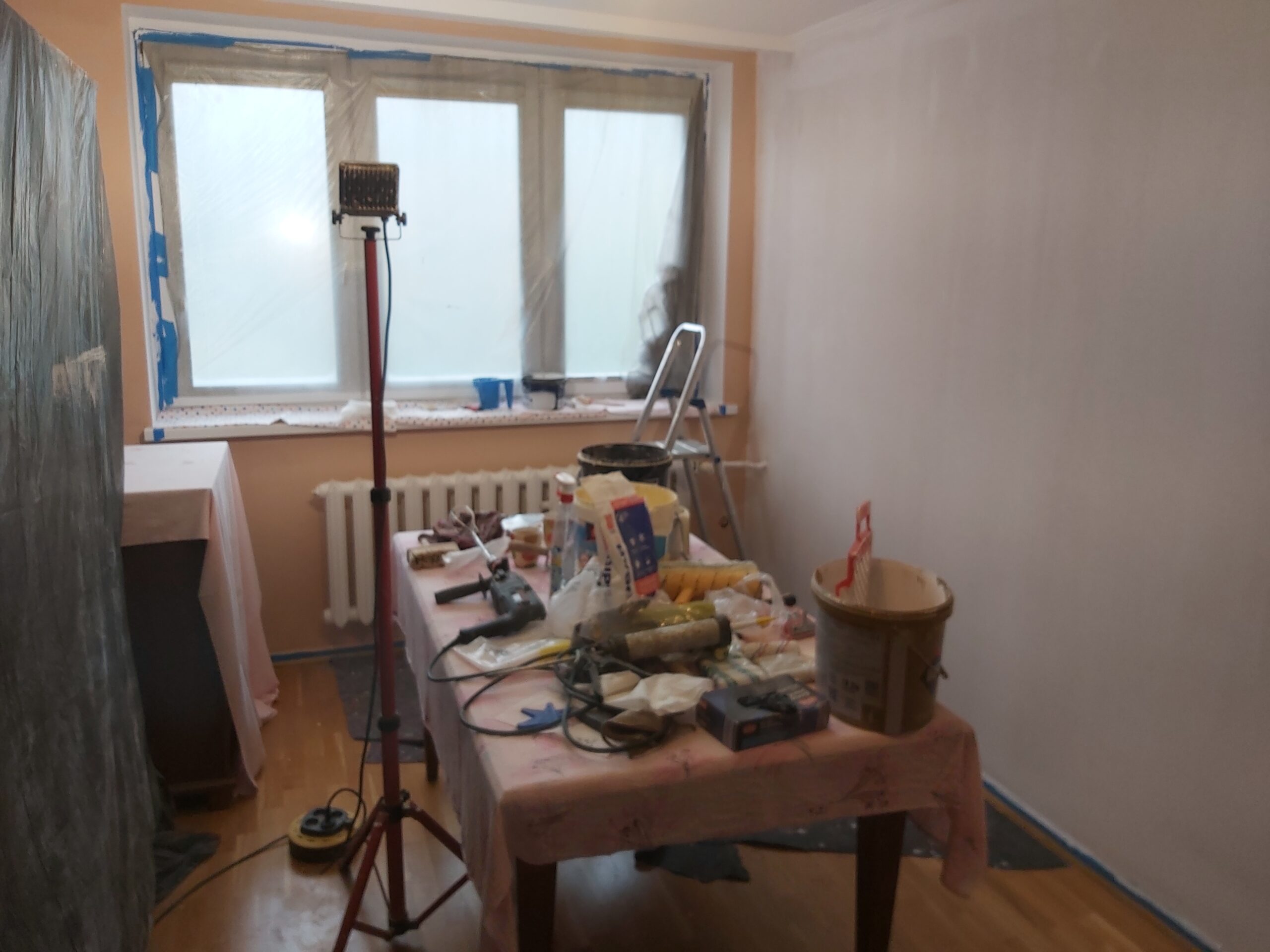 Pokój w trakcie remontu, z białą ścianą przygotowaną do malowania, stołem roboczym pełnym narzędzi i materiałów, lampą roboczą na statywie oraz drabiną przy oknie.