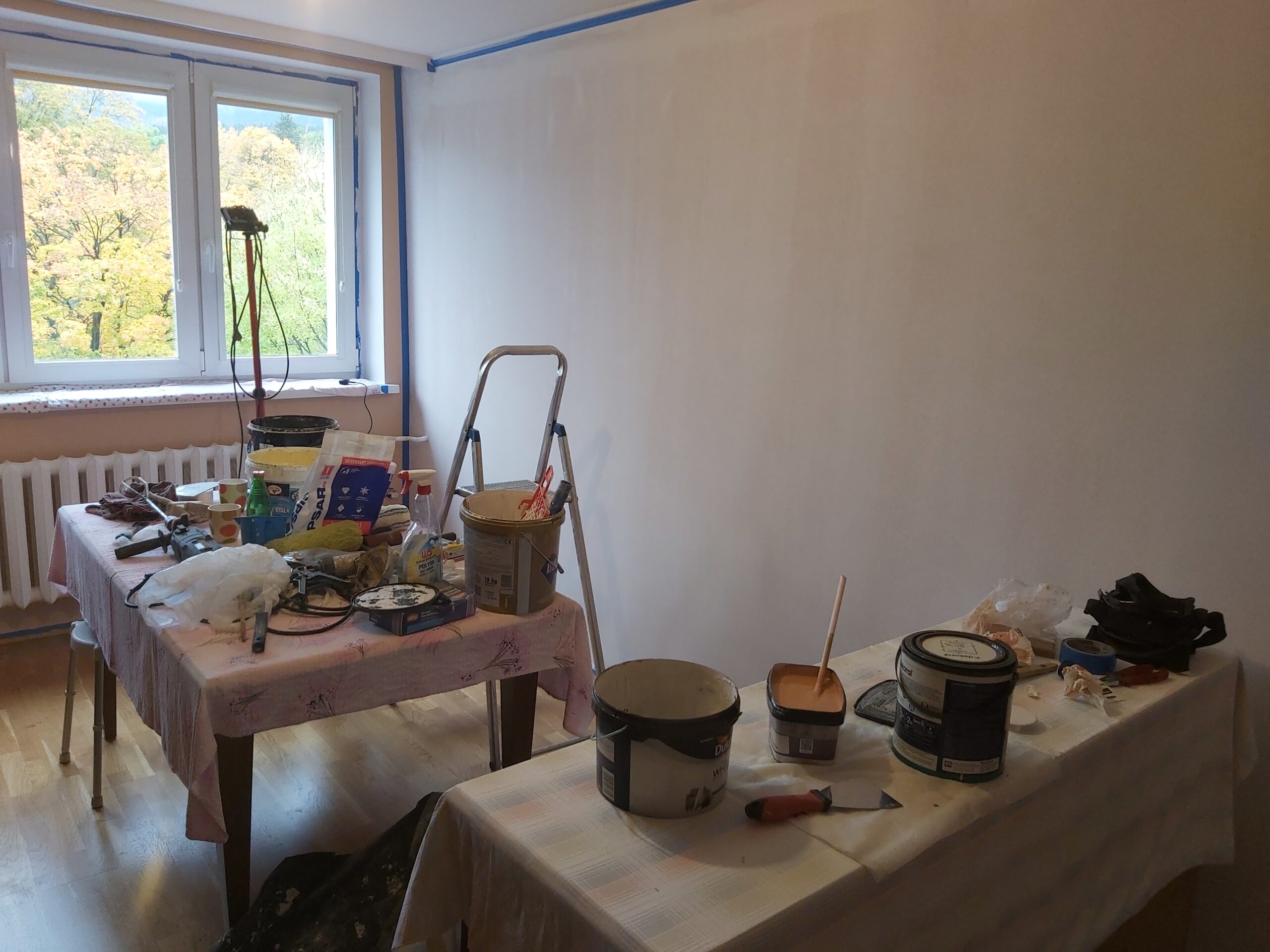 Pomieszczenie w trakcie prac wykończeniowych, z przygotowaną jasną ścianą, stołami przykrytymi foliami i farbami, narzędziami malarskimi oraz drabiną przy oknie.