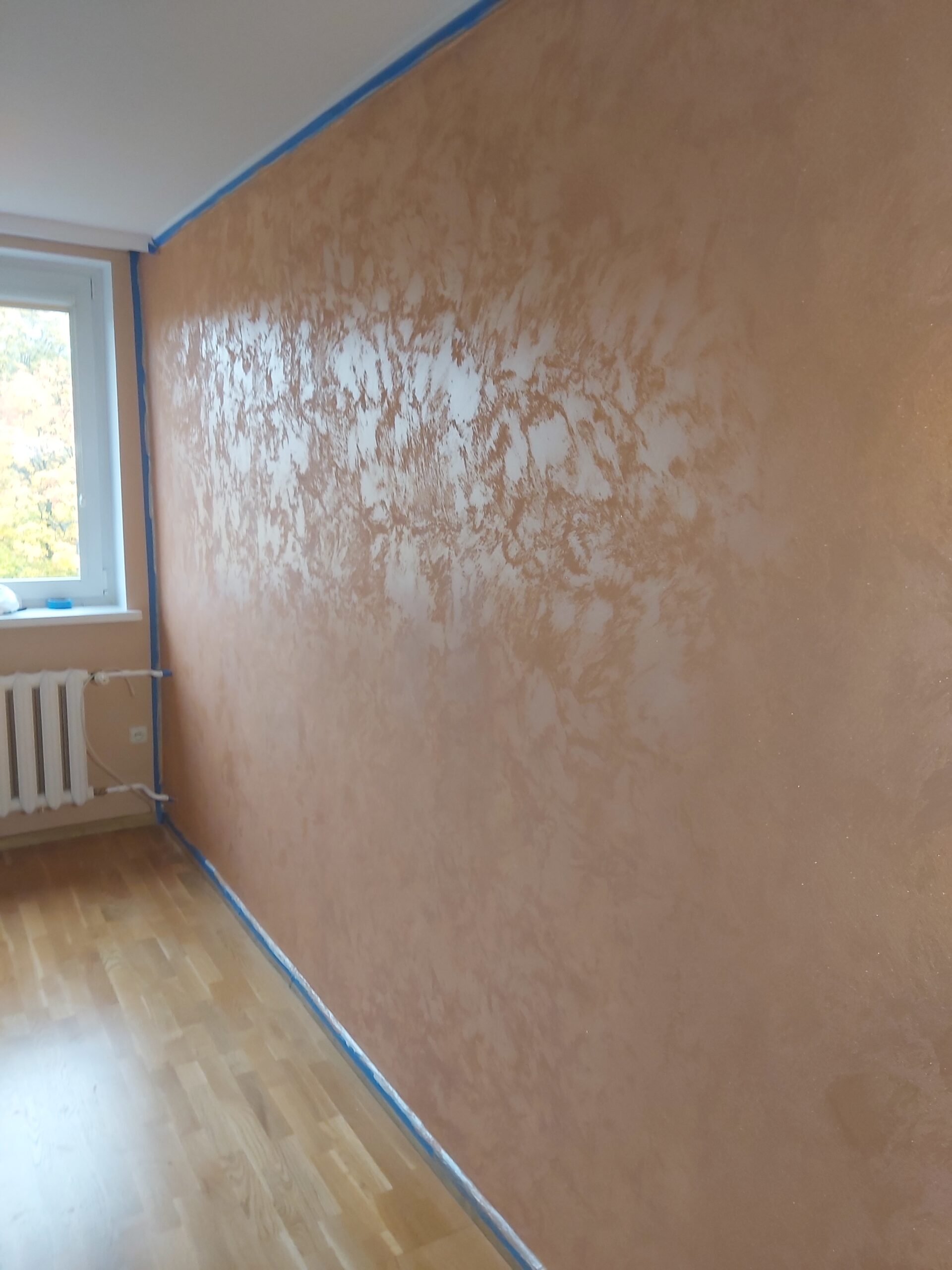 Ściana w pomieszczeniu wykończona dekoracyjną strukturą w ciepłym, brzoskwiniowo-beżowym kolorze, z widocznym połyskiem, zabezpieczona taśmą malarską przy suficie i podłodze, obok okna i grzejnika.