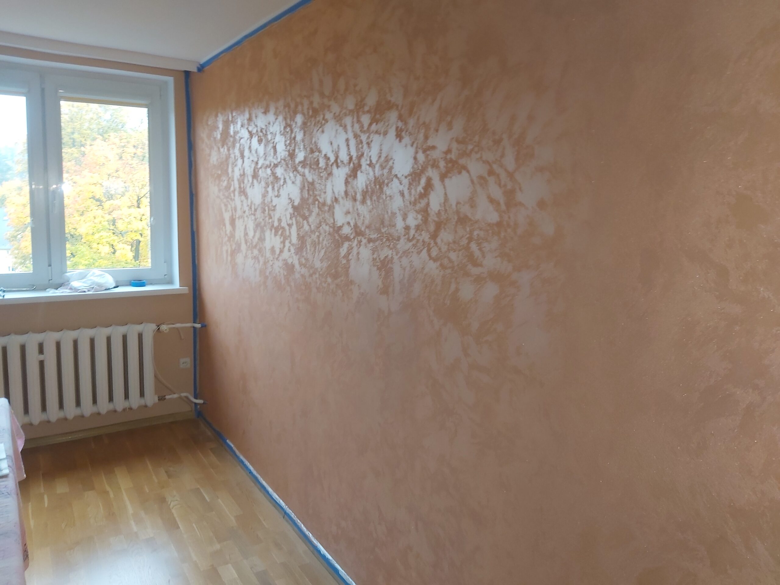 Ściana w pokoju wykończona dekoracyjną strukturą w ciepłym, beżowo-brzoskwiniowym odcieniu, z widocznym połyskiem, obok okna i grzejnika, zabezpieczona taśmą malarską przy krawędziach.