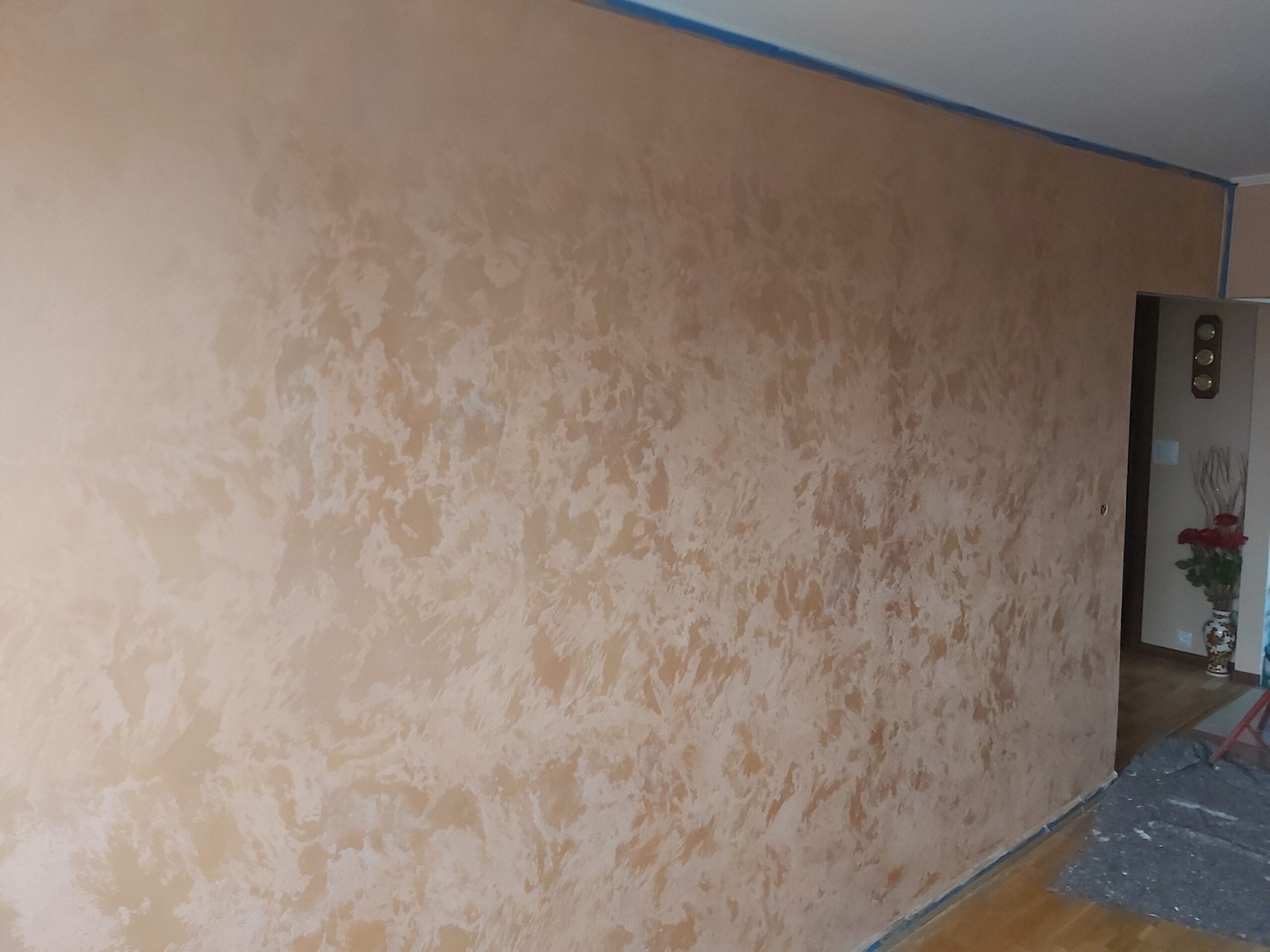 Długa ściana w pomieszczeniu wykończona dekoracyjną strukturą w jasnym, beżowo-brzoskwiniowym kolorze, zabezpieczona taśmą malarską przy krawędziach.