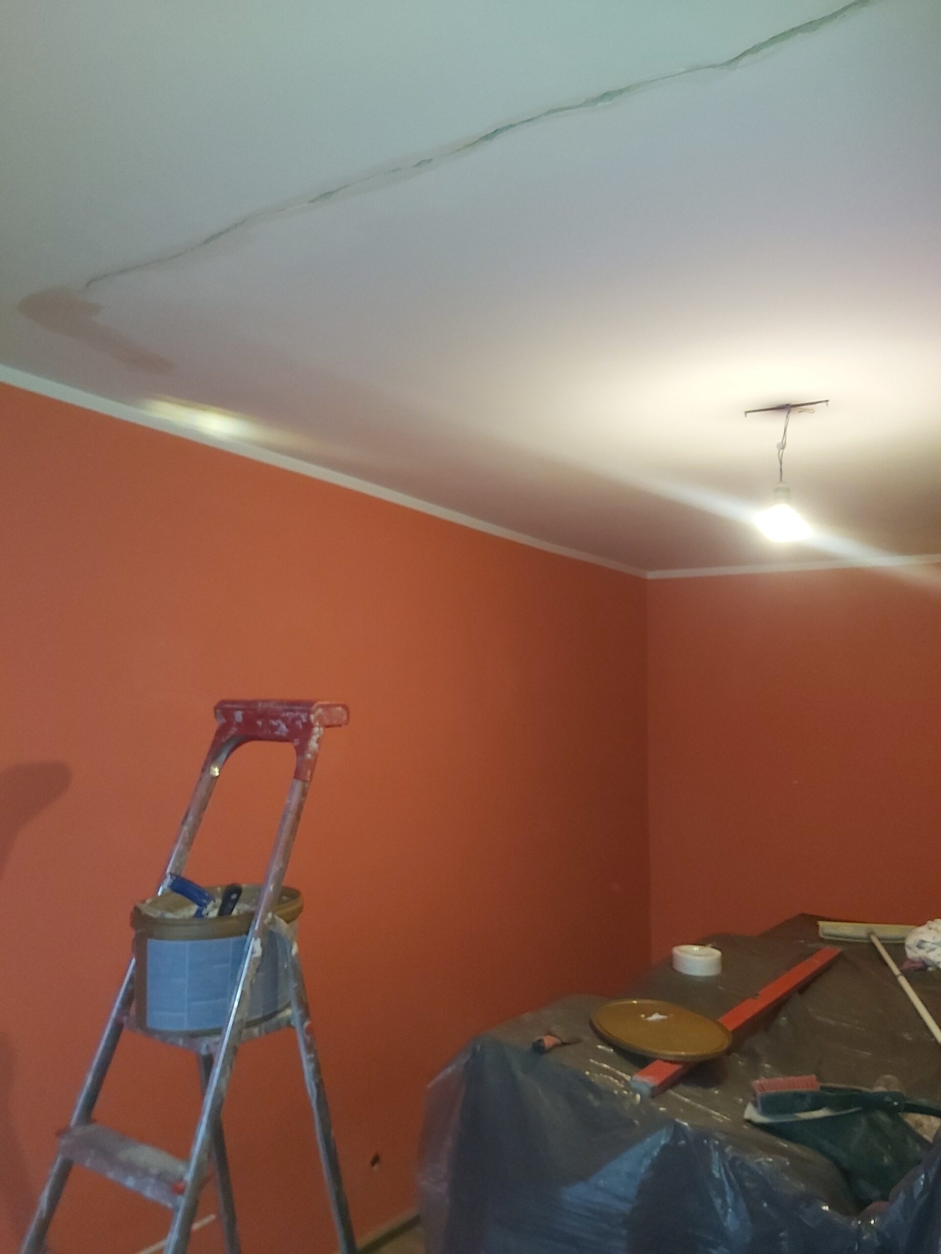 Pomieszczenie z pomarańczowymi ścianami w trakcie remontu, drabina malarska, zabezpieczone meble folią oraz sufit z widocznymi poprawkami i pęknięciem przy krawędzi.