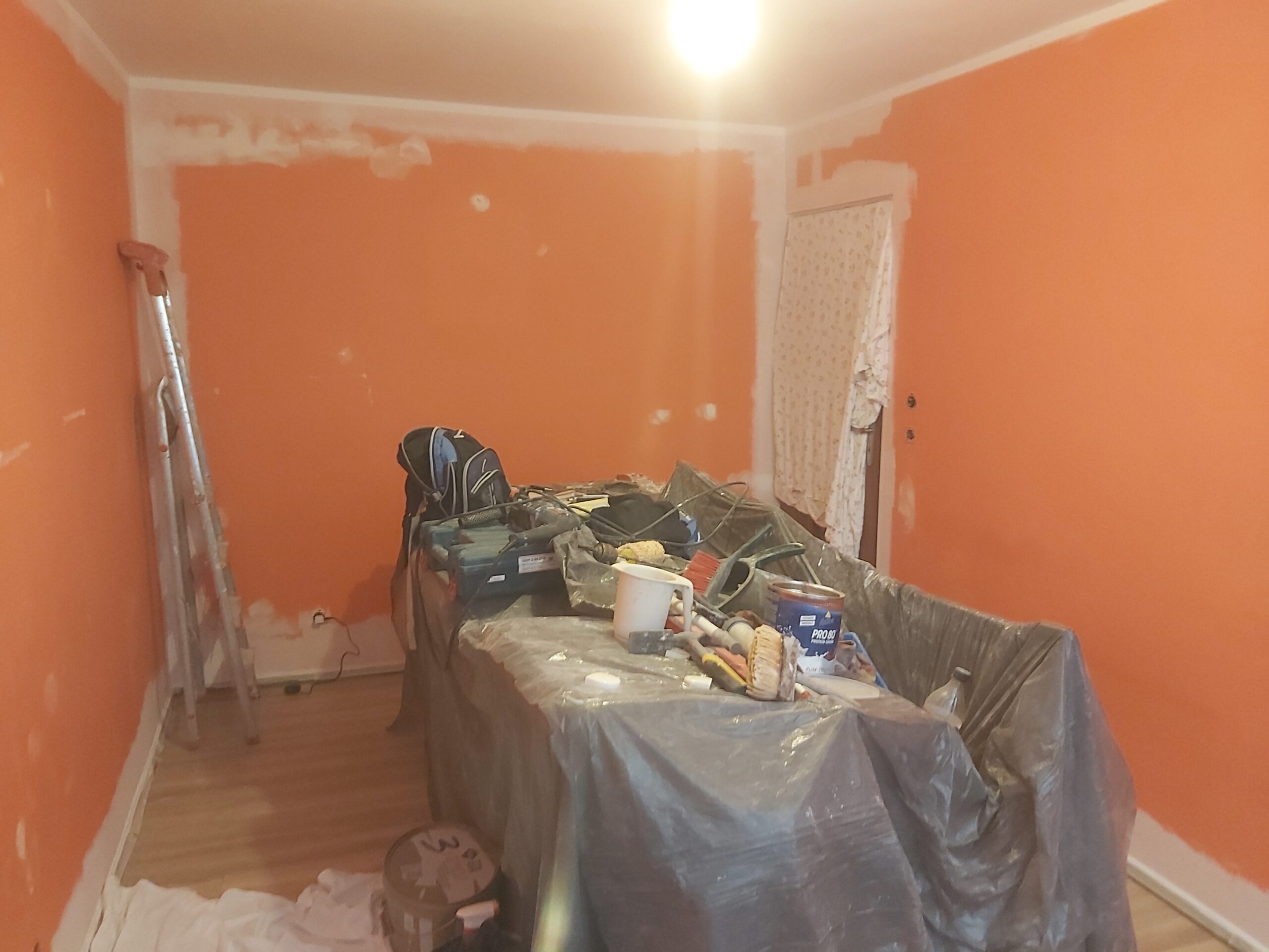 Wnętrze pokoju w trakcie remontu z pomalowanymi na pomarańczowo ścianami, zabezpieczonymi meblami oraz narzędziami i materiałami malarskimi.