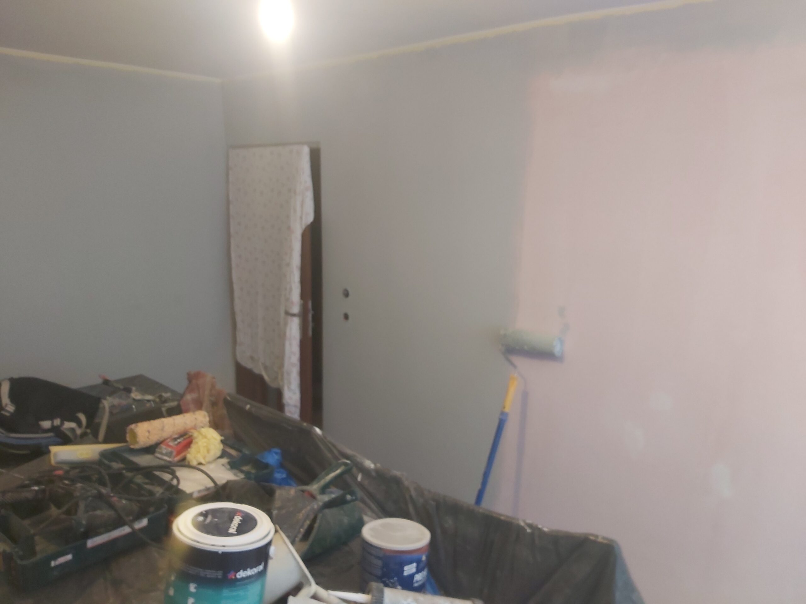 Ściana w trakcie malowania wałkiem, widoczne fragmenty świeżej farby, zabezpieczone meble i narzędzia malarskie w pomieszczeniu.