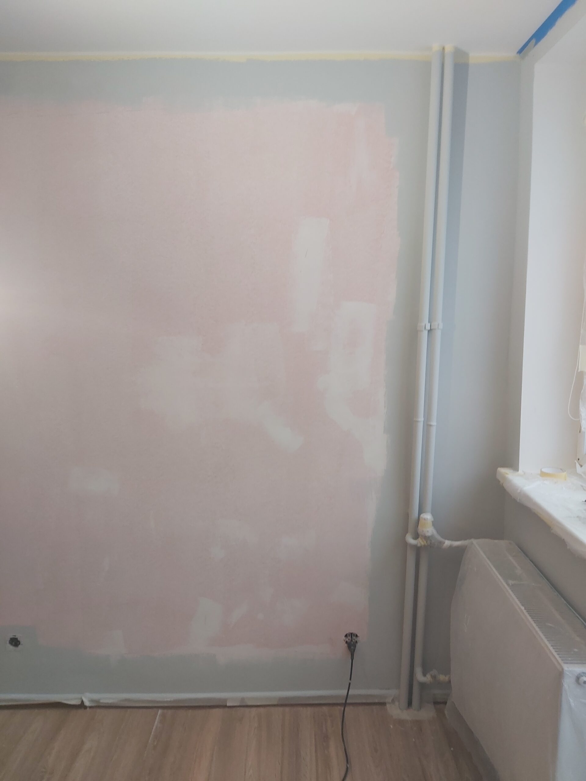 Ściana w trakcie przygotowania do malowania, widoczne miejsca po szpachlowaniu, rury instalacyjne przy narożniku oraz zabezpieczony grzejnik i parapet.