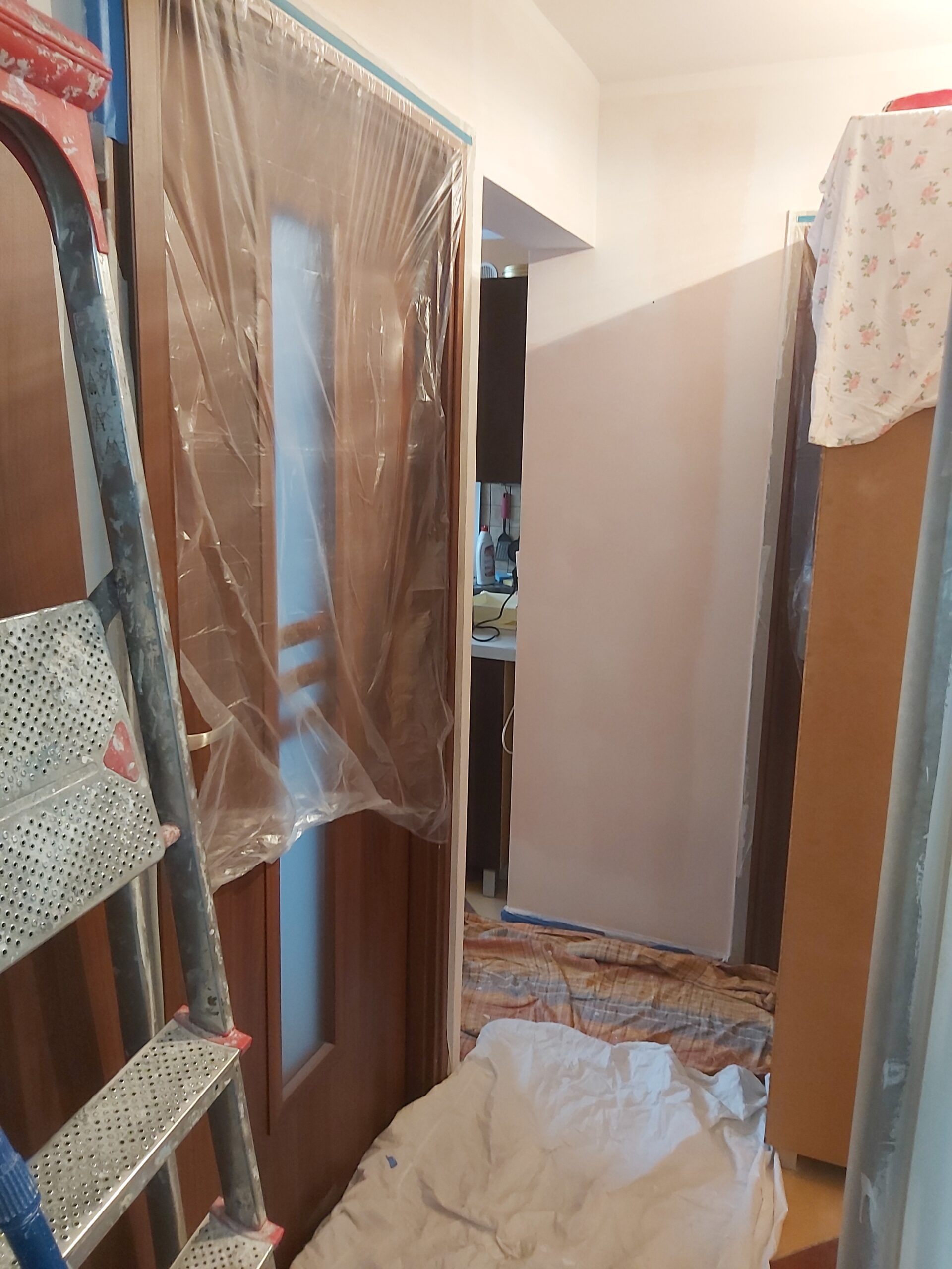 Wąski korytarz w trakcie malowania, ściana pokryta jasną farbą, drzwi zabezpieczone folią ochronną, drabina malarska i zabezpieczenia na podłodze.