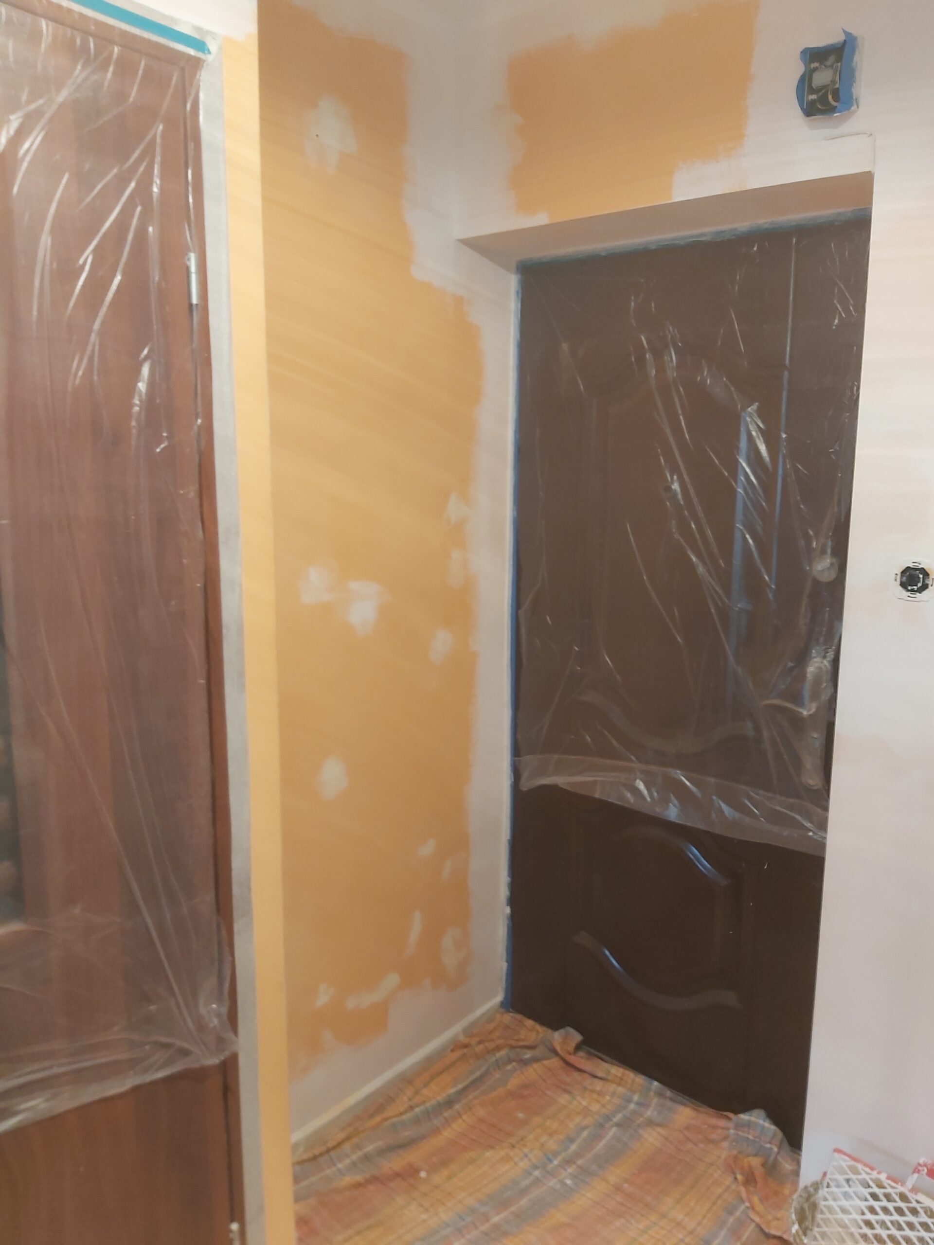 Ściana w przedpokoju w trakcie przygotowania do malowania, fragmenty pokryte farbą podkładową, drzwi zabezpieczone folią malarską, taśma ochronna przy krawędziach.