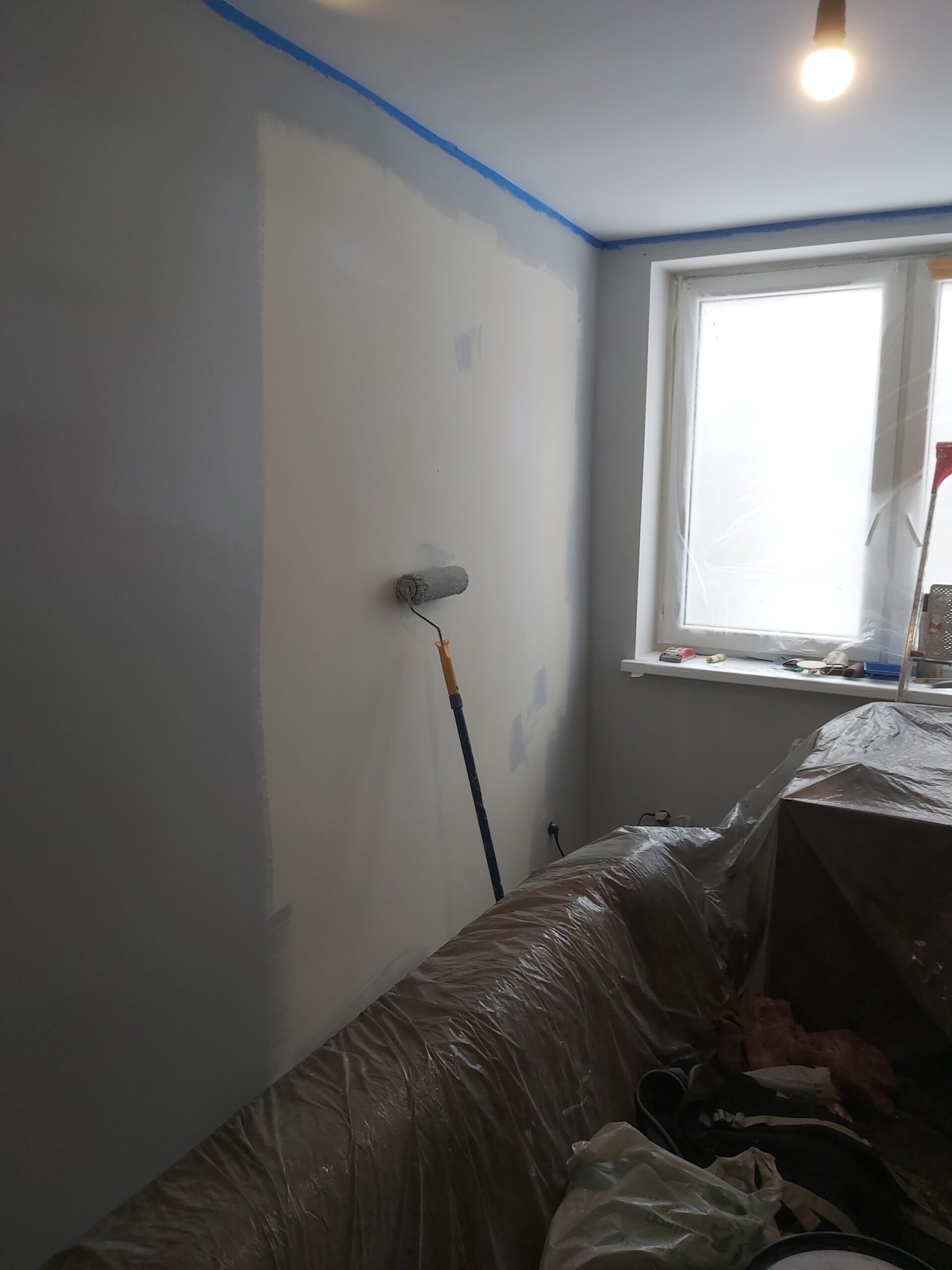 Ściana w trakcie malowania jasną farbą, wałek oparty o powierzchnię ściany, okno zabezpieczone folią i taśmą malarską, meble przykryte folią ochronną.