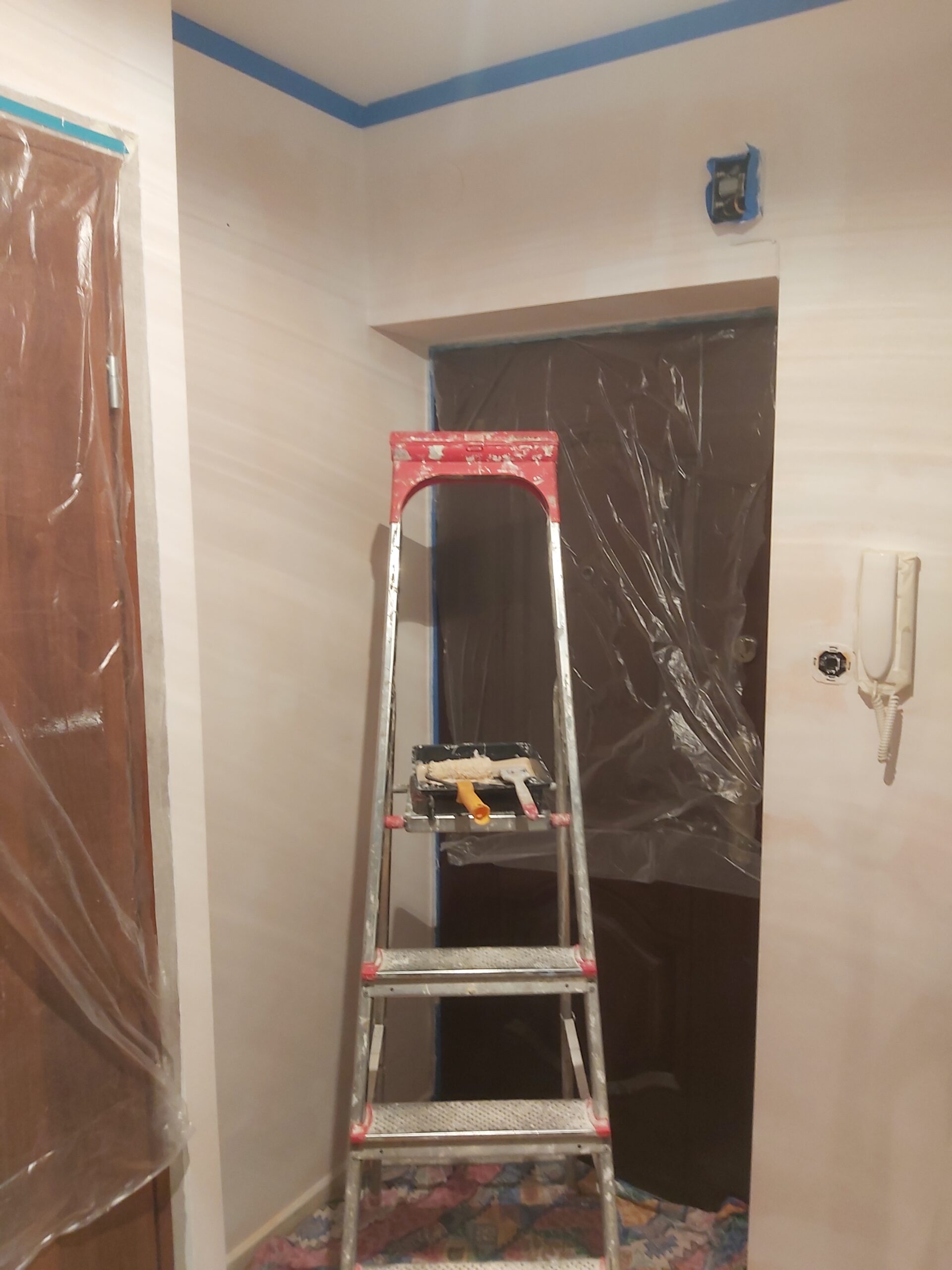 Drabina ustawiona w przedpokoju przed drzwiami zabezpieczonymi folią, ściany w trakcie malowania z użyciem taśmy ochronnej i widocznymi narzędziami malarskimi.