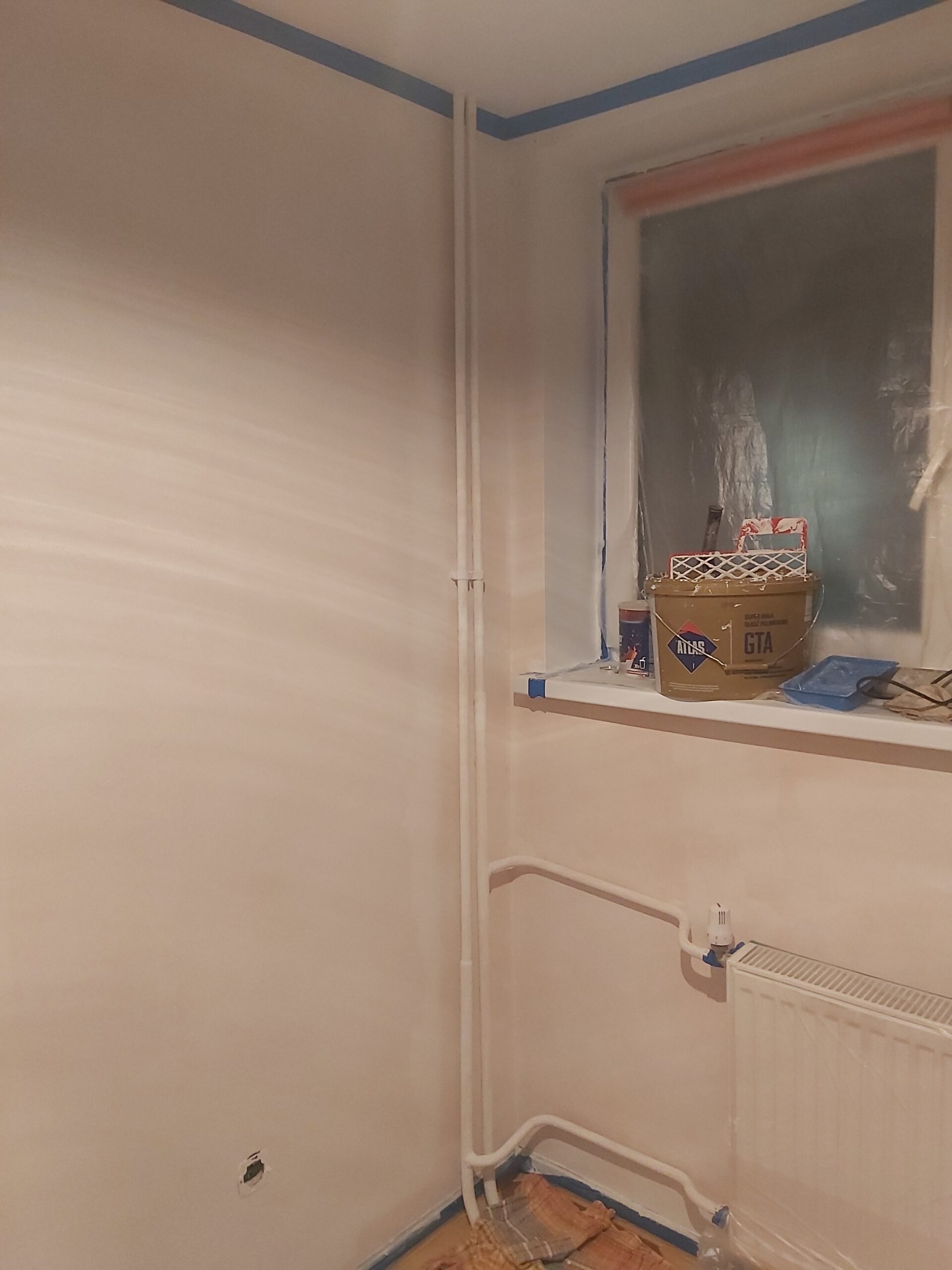 Róg pokoju z oknem zabezpieczonym folią, widocznymi rurami grzewczymi i kaloryferem, ściany w trakcie malowania z użyciem taśmy ochronnej.