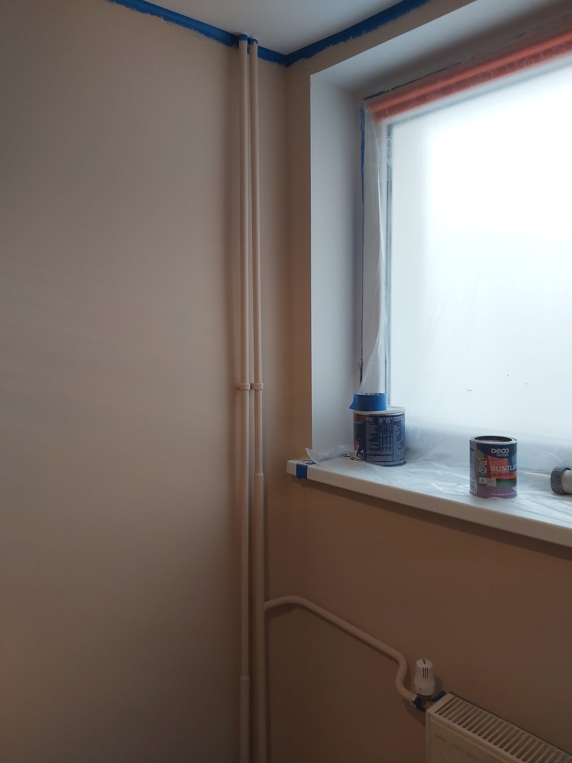 Róg pokoju z oknem, widocznymi pionowymi rurami grzewczymi oraz parapetem zabezpieczonym folią i taśmą malarską podczas prac wykończeniowych.
