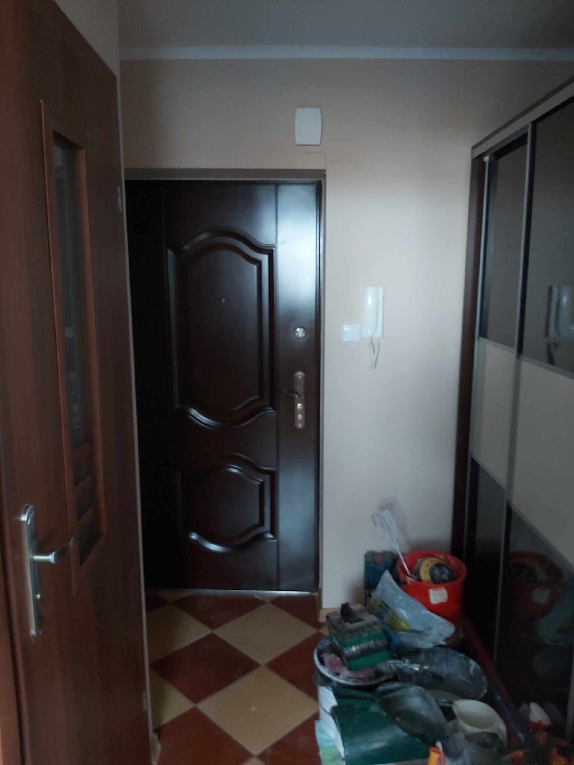 Przedpokój z zamontowanymi ciemnymi drzwiami wejściowymi, jasnymi ścianami w ciepłym odcieniu oraz widocznymi narzędziami i materiałami pozostałymi po pracach remontowych.