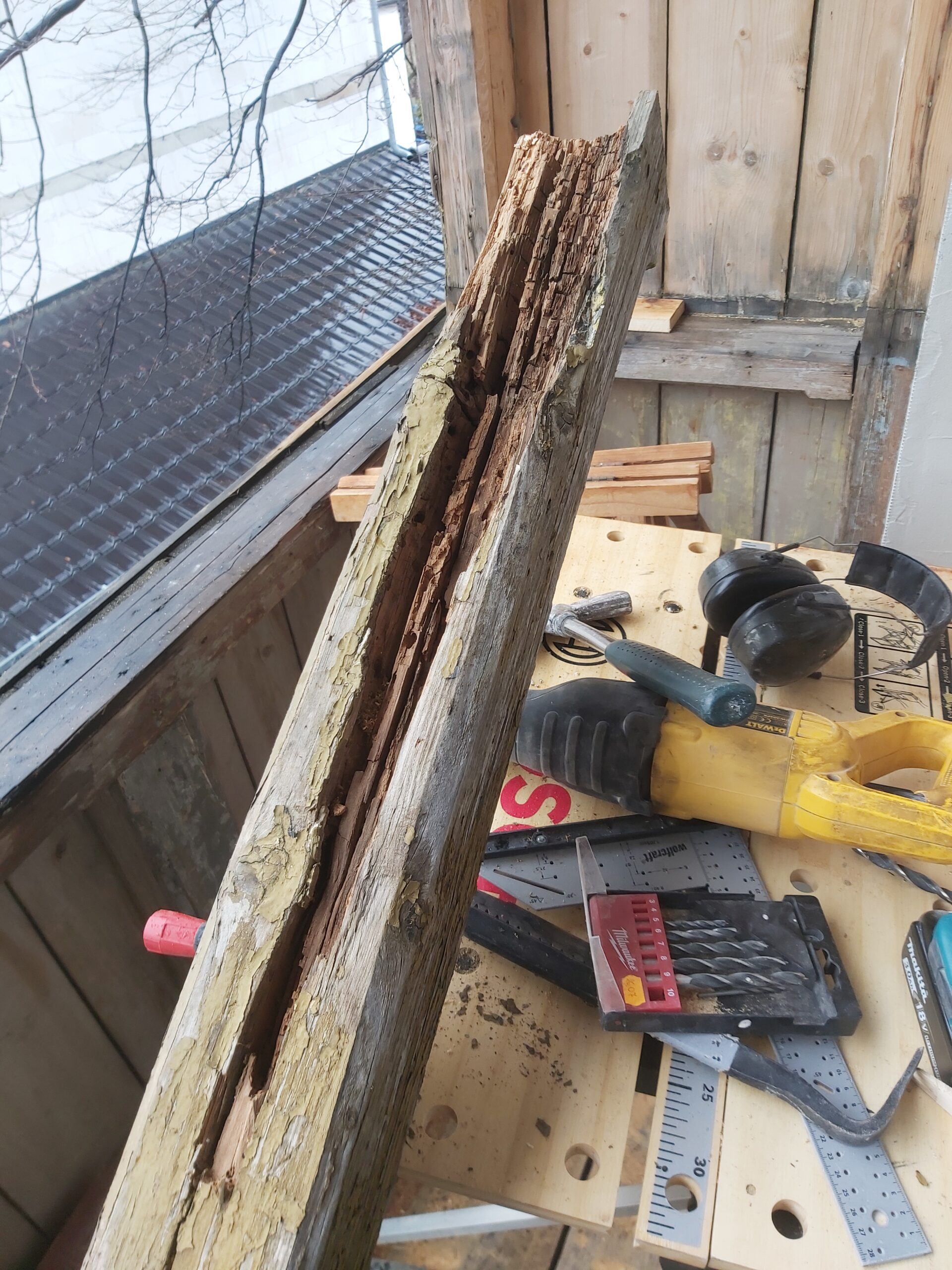 Zniszczony, spróchniały drewniany element balkonu z widocznymi ubytkami drewna, ułożony na stole warsztatowym podczas prac remontowych.