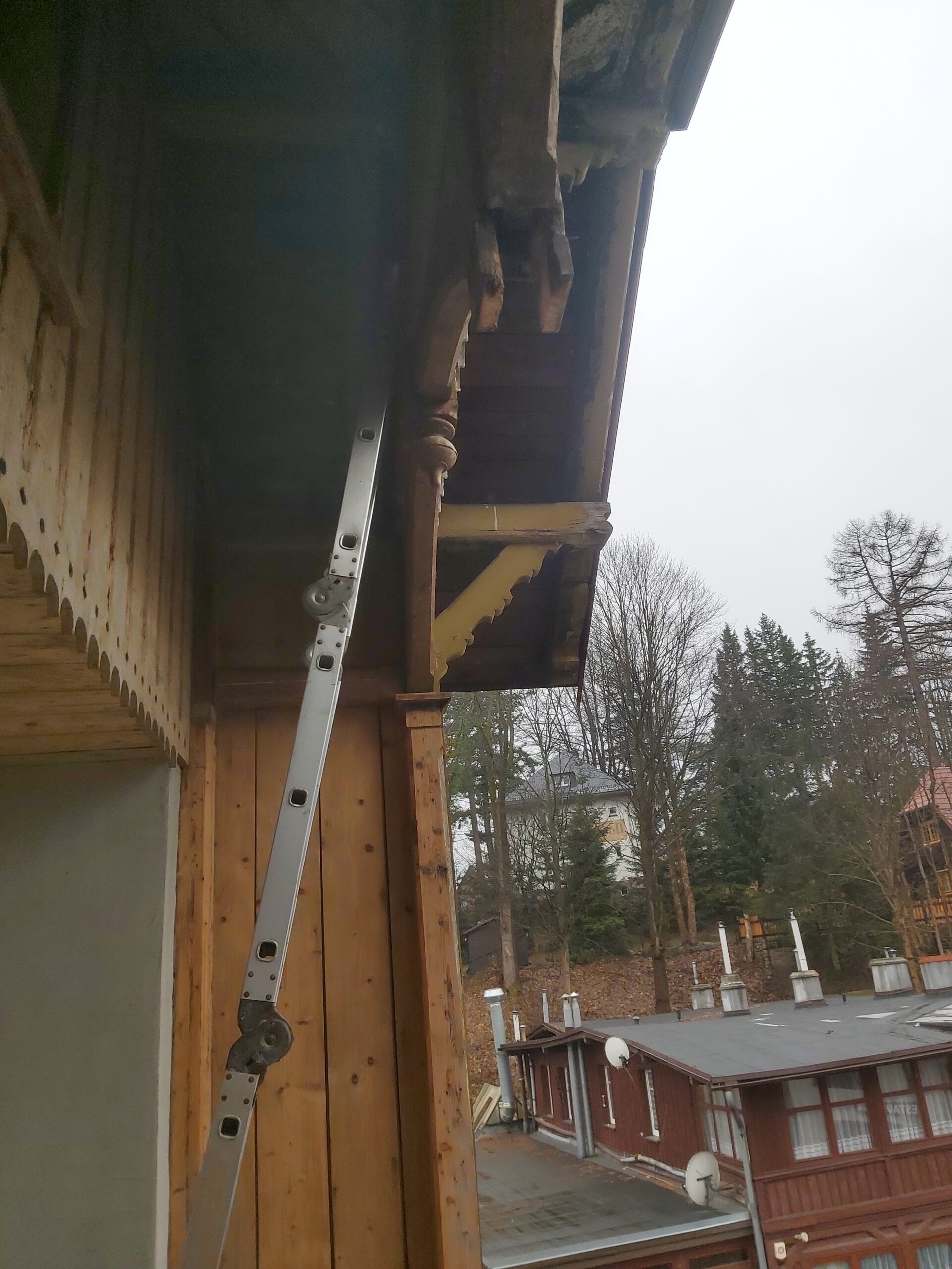 Fragment drewnianej konstrukcji balkonu z ozdobnym słupem i elementami dachu, widoczna drabina oraz ślady prac remontowych.