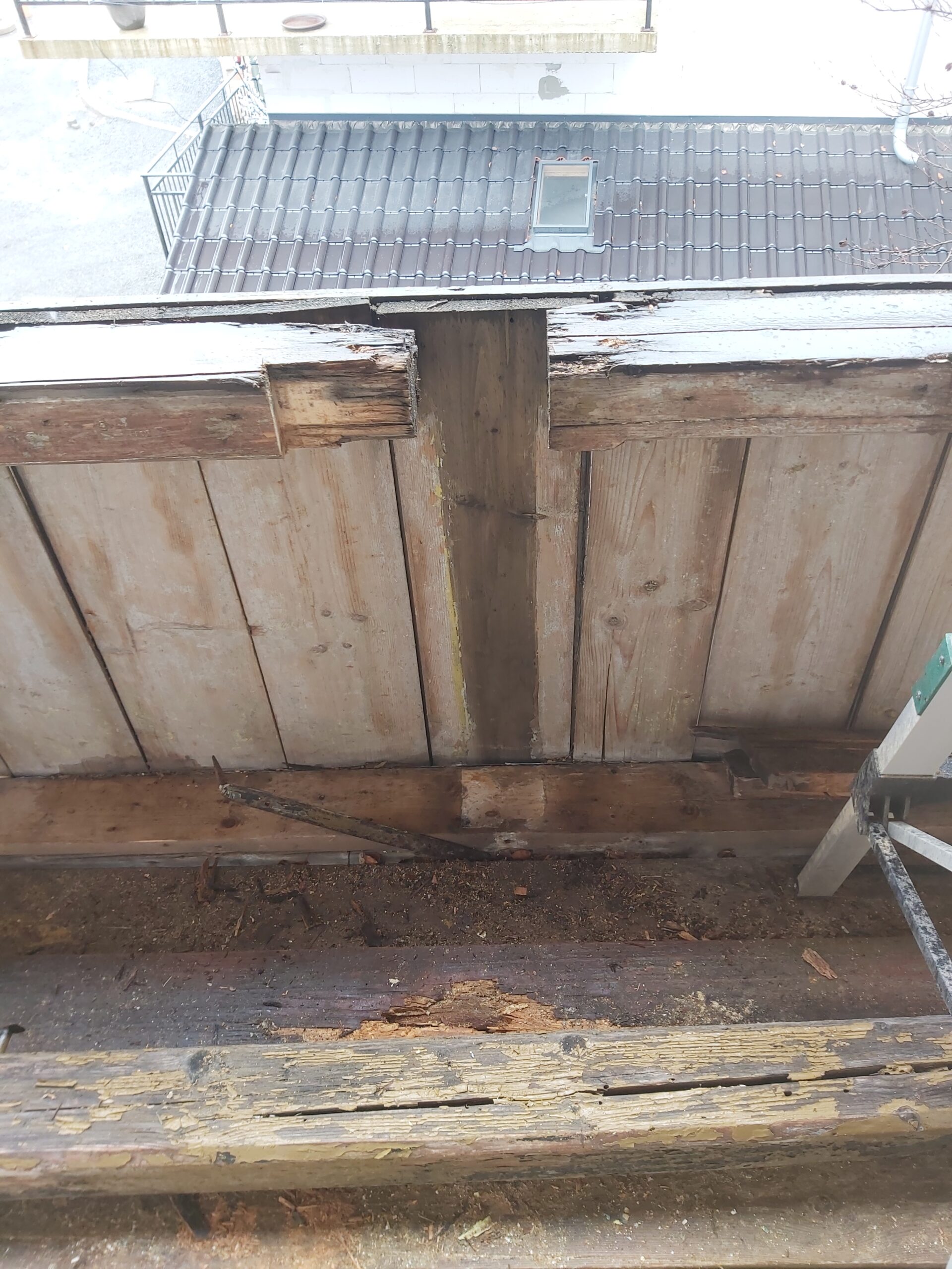 Widok na zniszczone elementy drewnianej konstrukcji balkonu z widoczną korozją biologiczną i ubytkami drewna przy krawędzi podłogi.