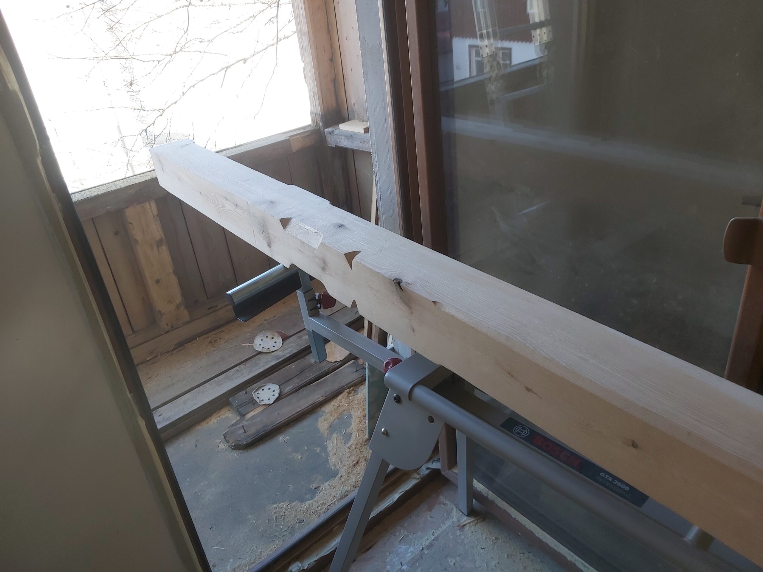 Drewniana belka z precyzyjnie wykonanymi wycięciami konstrukcyjnymi, ułożona na stojaku roboczym podczas prac stolarskich przy oknie.