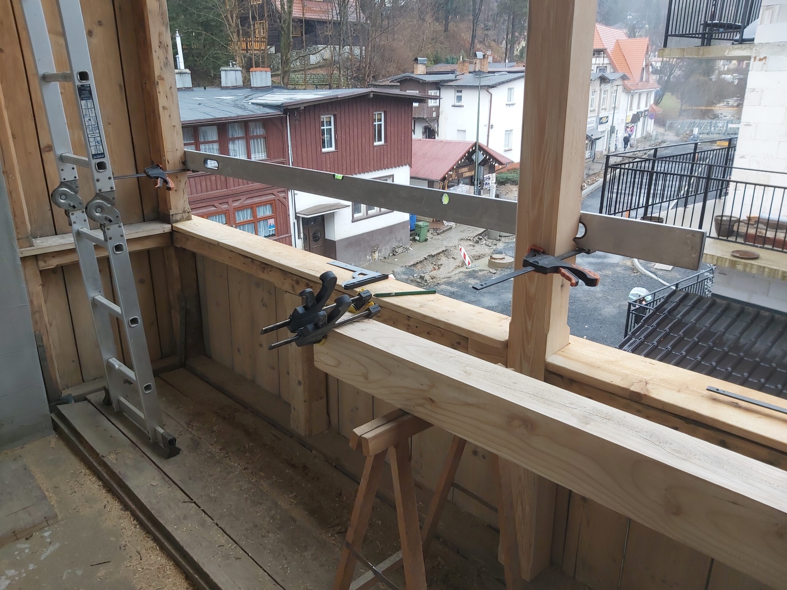 Drewniany balkon w trakcie remontu, z elementami konstrukcyjnymi zamocowanymi ściskami stolarskimi oraz widokiem na okoliczne budynki i ulicę.