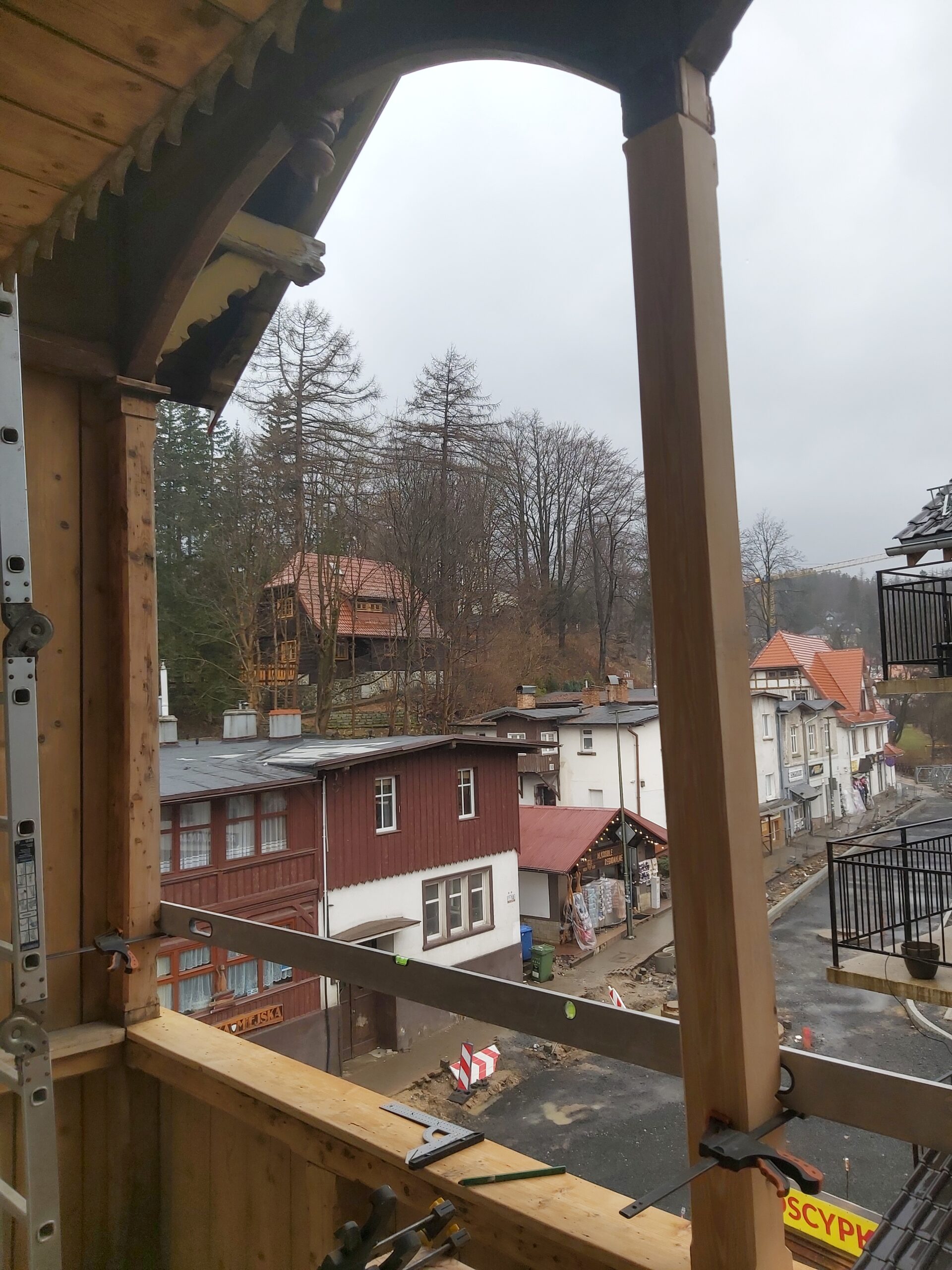 Widok z drewnianego balkonu w trakcie remontu, z zamontowanymi elementami konstrukcyjnymi i panoramą okolicznych budynków oraz ulicy.