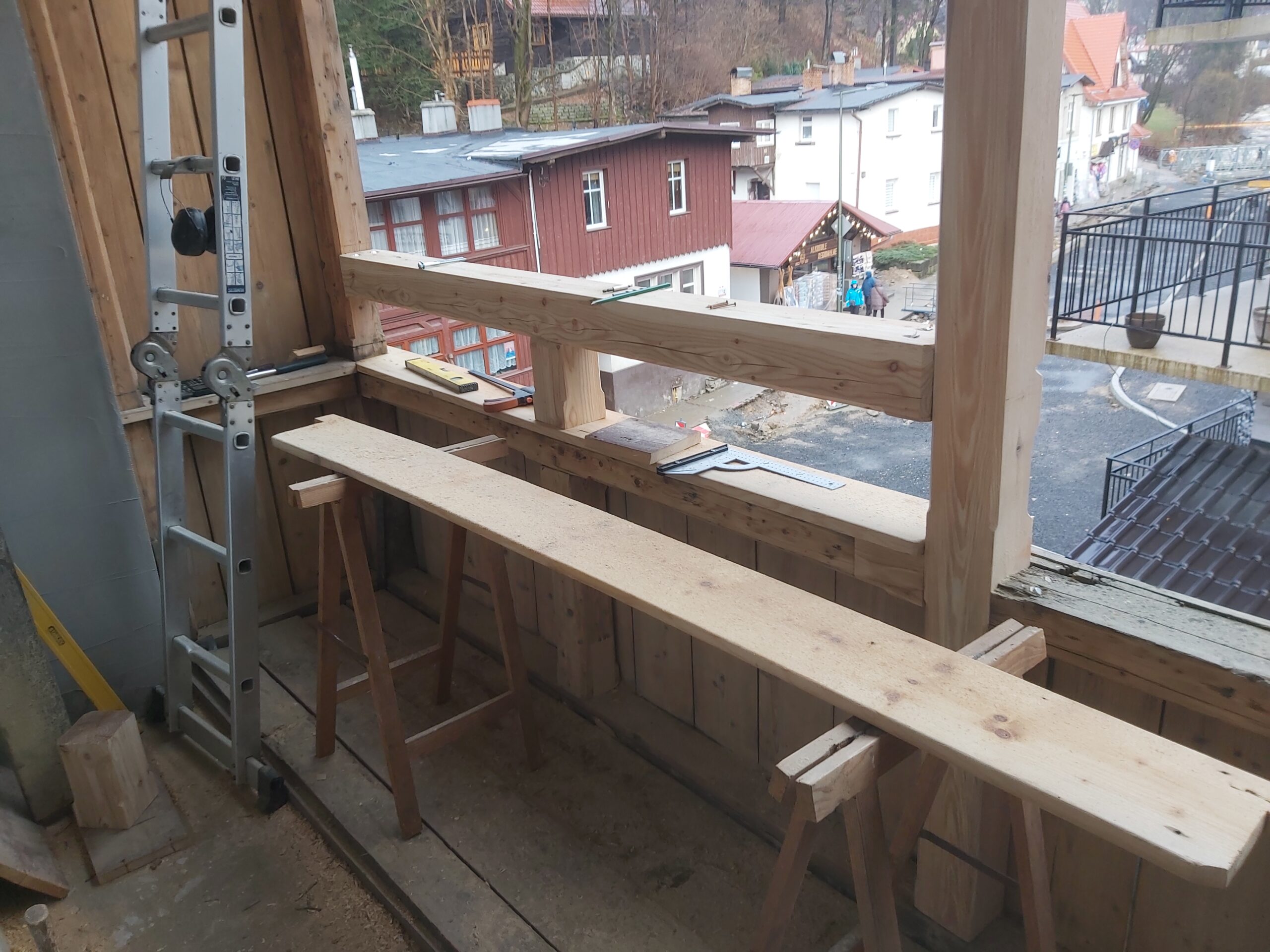 Drewniane deski ułożone na kozłach roboczych na balkonie w trakcie prac stolarskich, w tle widok na zabudowania.