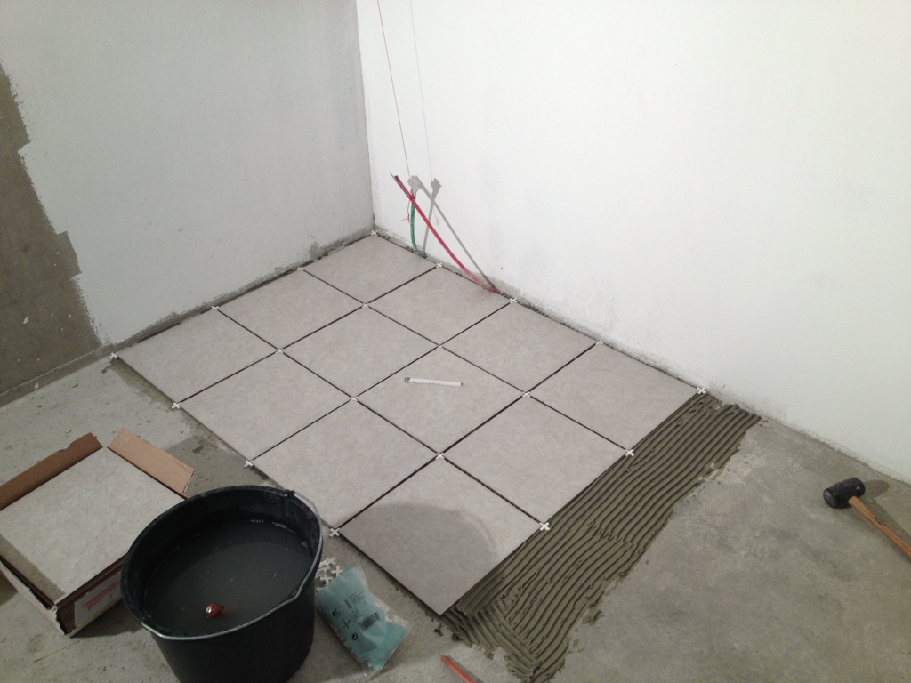 Kwadratowe płytki podłogowe układane przy ścianie, widoczny klej do płytek oraz narzędzia podczas prac wykończeniowych.