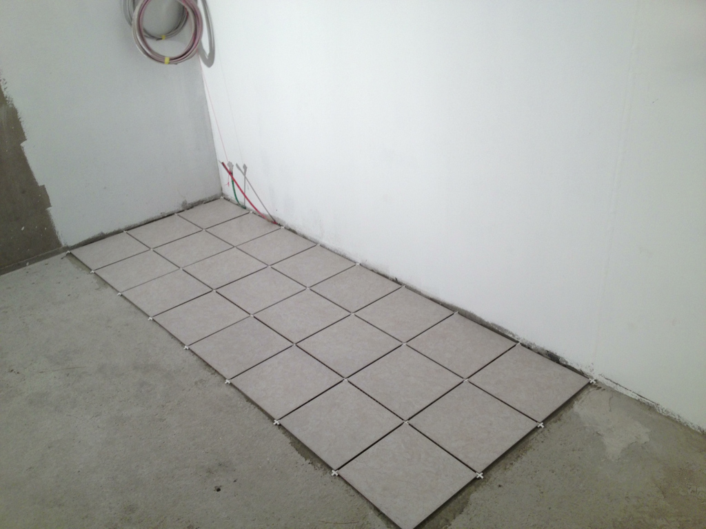 Kwadratowe płytki podłogowe ułożone przy ścianie w narożniku pomieszczenia, widoczny etap prac wykończeniowych.