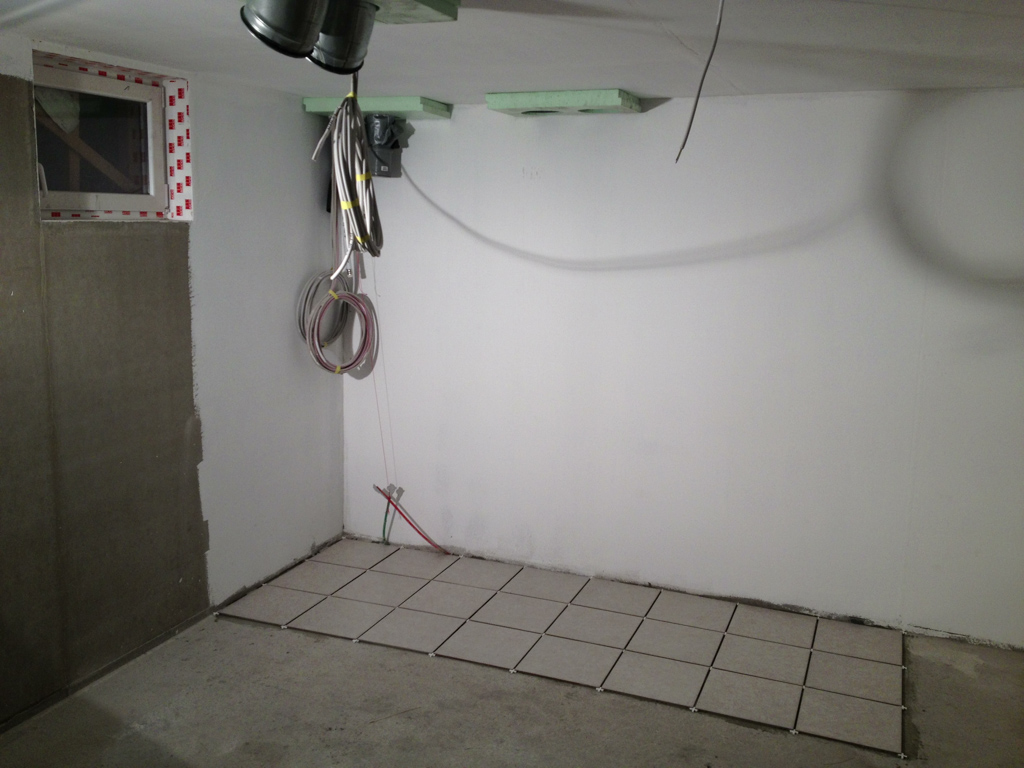 Szare płytki podłogowe ułożone przy ścianie technicznej z widoczną instalacją i przewodami, etap prac wykończeniowych.