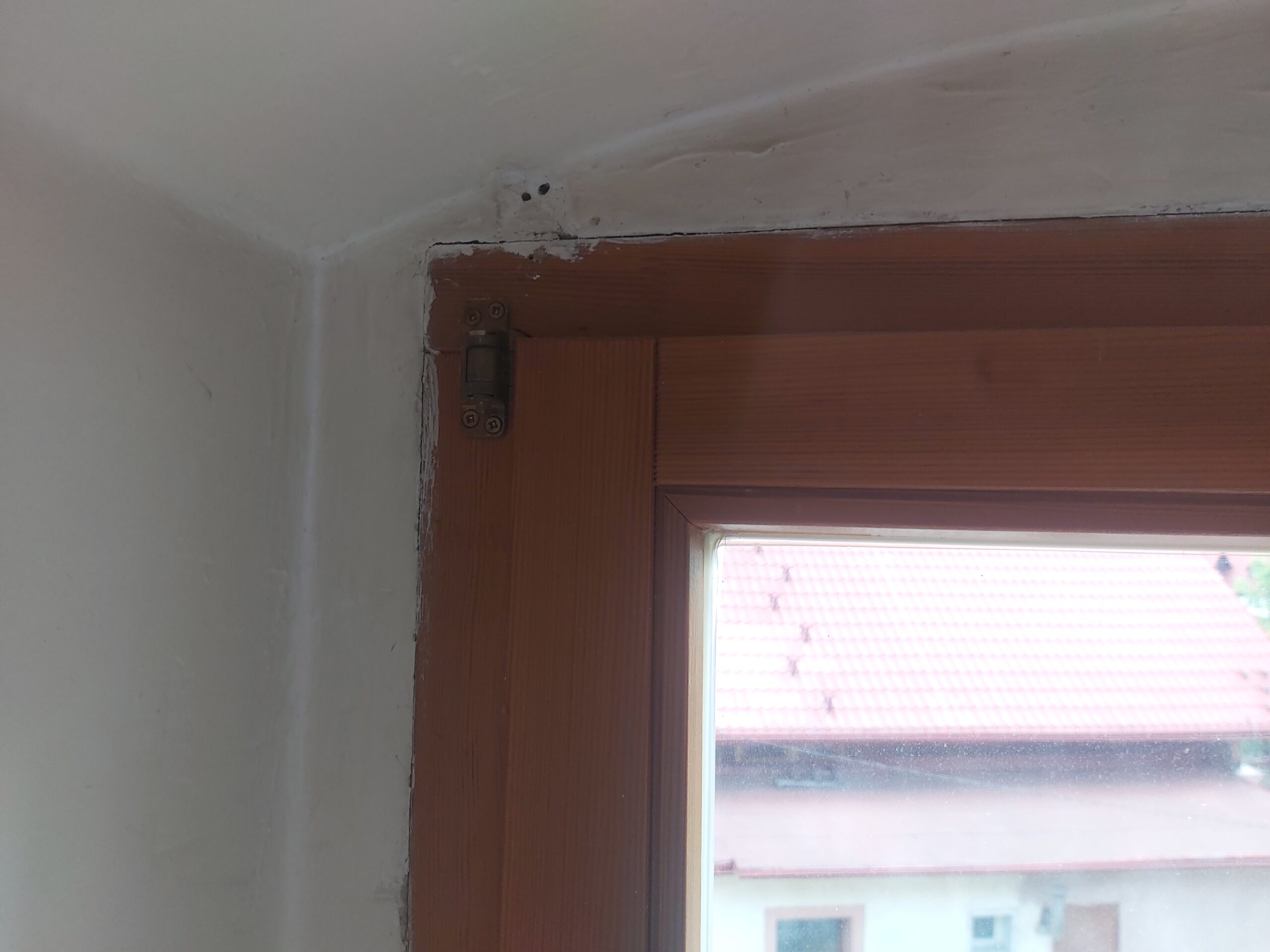 Zbliżenie na górny narożnik drewnianej ramy okiennej przy ścianie, widoczny zawias, krawędź tynku oraz sufit po pracach remontowych.