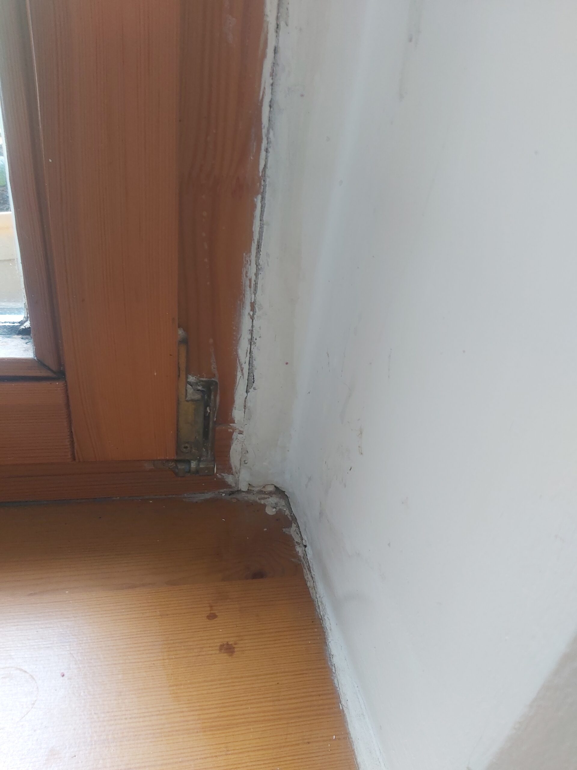 Zbliżenie na dolny róg ramy okiennej i ścianę po pracach remontowych, widoczne uzupełnienia tynku i miejsce przy zawiasie okna.