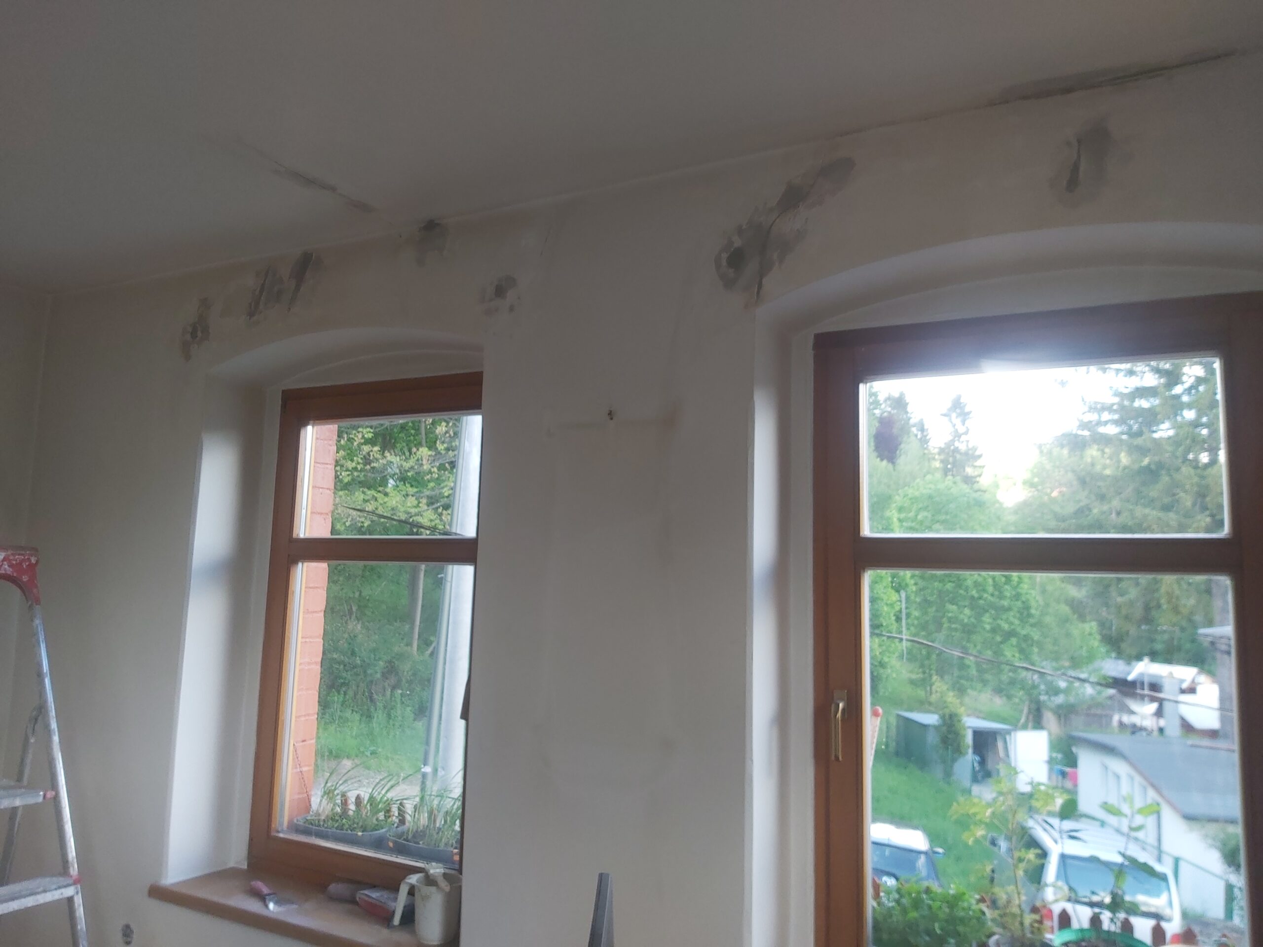 Ściana z dwoma oknami w trakcie prac wykończeniowych, widoczne miejsca napraw i szpachlowania nad nadprożami przed malowaniem.
